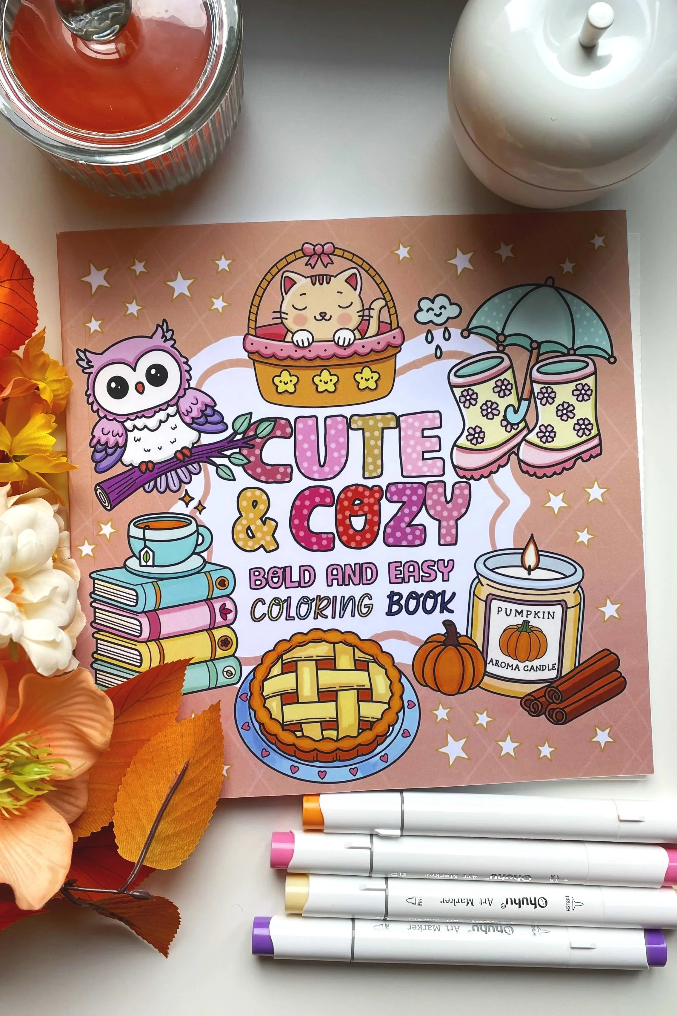 Coloring book titled "Cute & Cozy" with illustrations of a cat in a basket, an owl, rain boots, books, a pumpkin pie, a pumpkin, and cinnamon sticks, surrounded by fall leaves and artificial flowers. There are six colorful markers placed below the book.