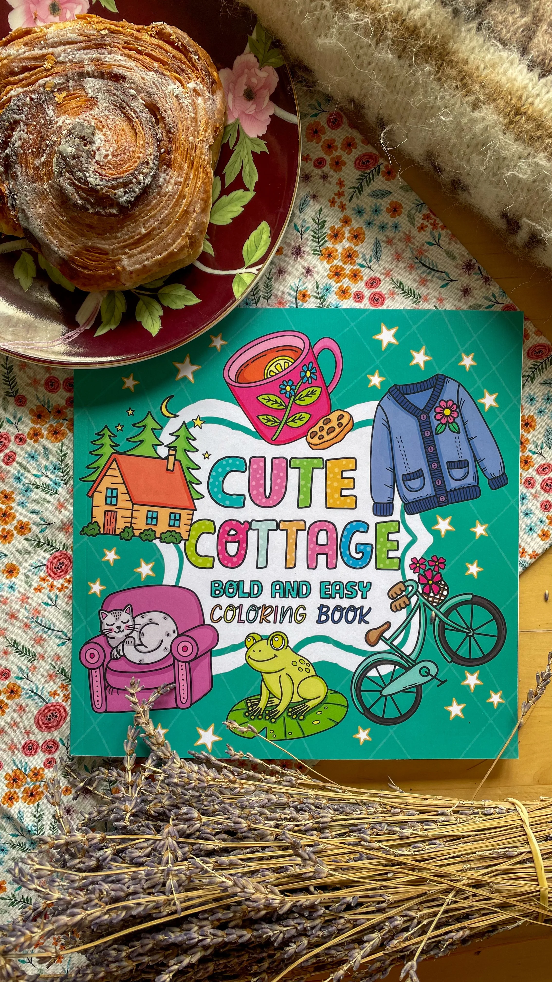 A colorful outdoor scene with a book titled 'Cute Cottage' coloring book, a plate with a flaky croissant, and a bunch of dried lavender. The scene is set on a floral patterned cloth with some knitted fabric in the background.