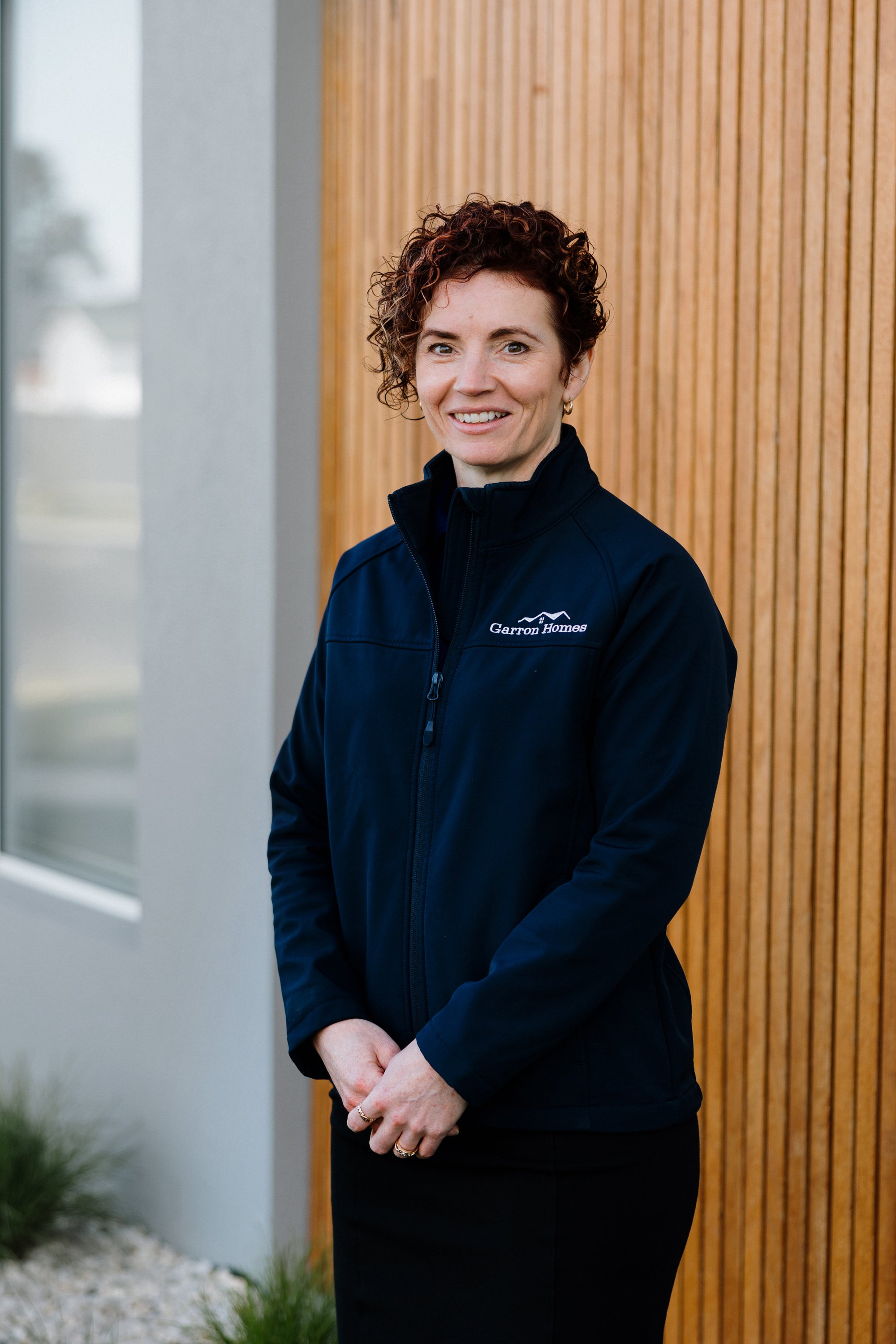 Portrait of a woman with short curly hair, wearing a black zip-up jacket with the logo "Garrol Homes," standing outside near a wooden wall and a window, smiling at the camera.