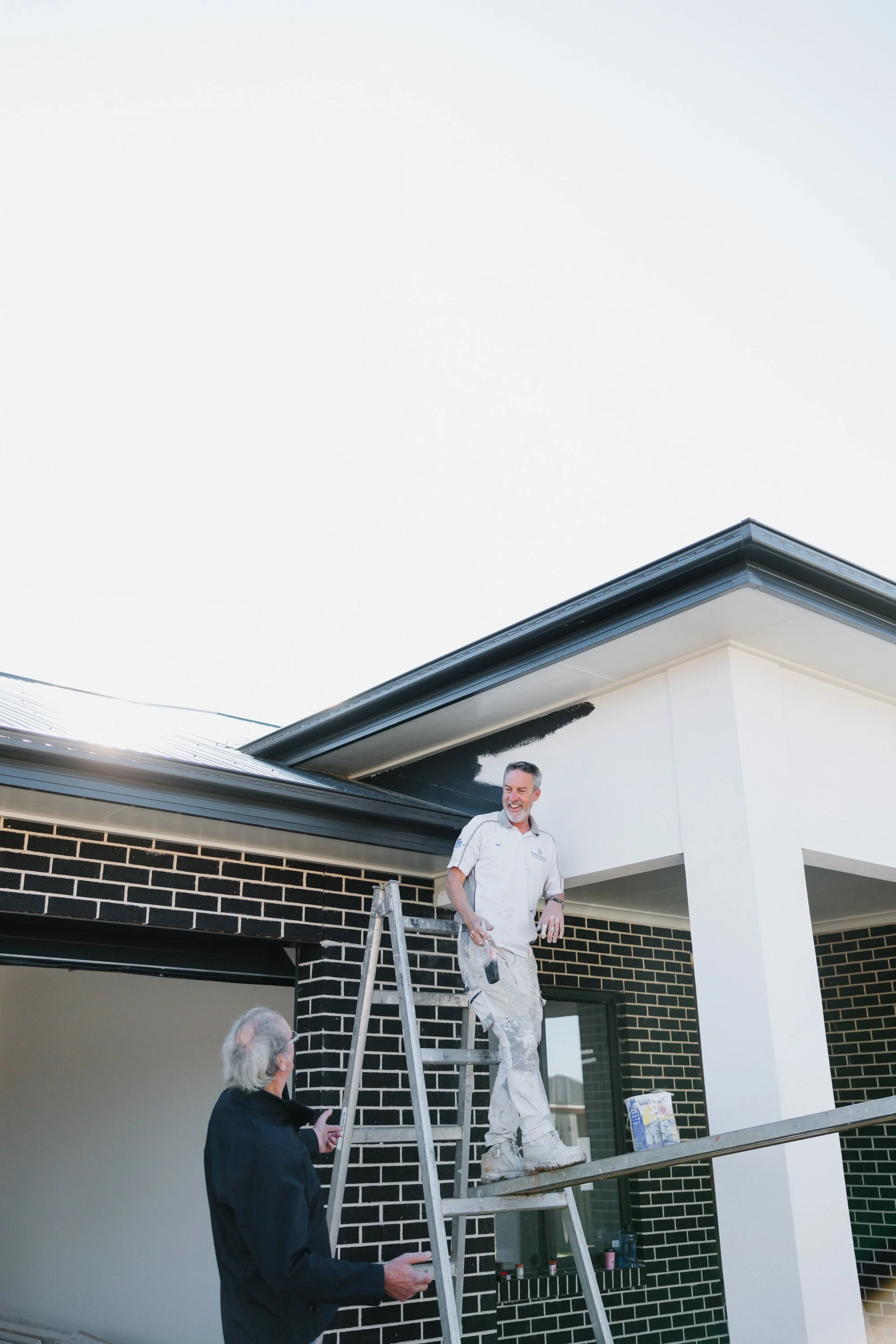 A man in white painter's clothing standing on a ladder, holding a paint roller, painting the exterior of a house. Another man with white hair and a beard, dressed in black, is standing below, and a third man on a platform beneath them is assisting. The house has black brick on lower walls and white walls above, with a porch area.