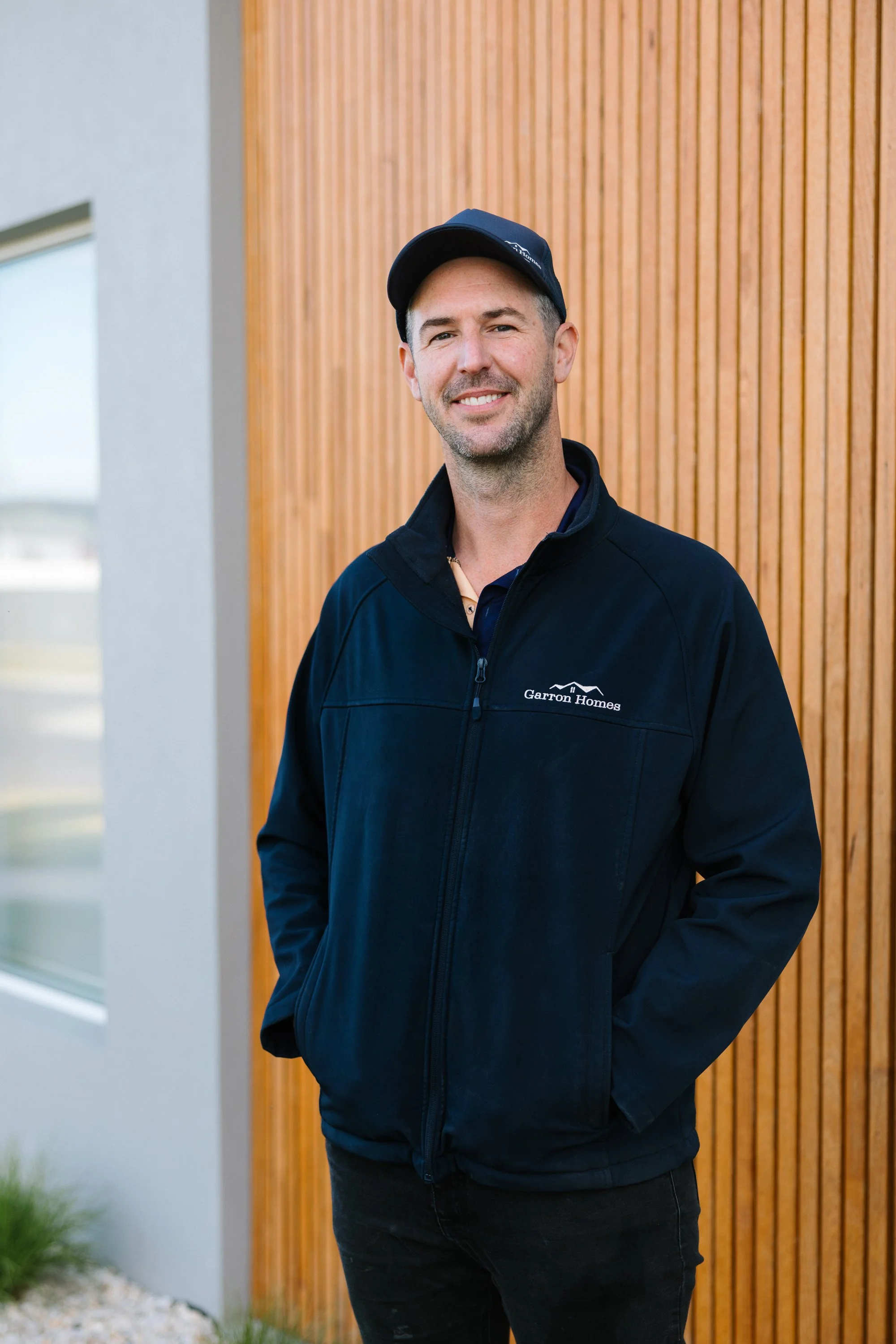 A man standing outside in front of a modern building with vertical wooden siding, wearing a black jacket with 'Garron Homes' logo, a navy cap, and smiling at the camera.