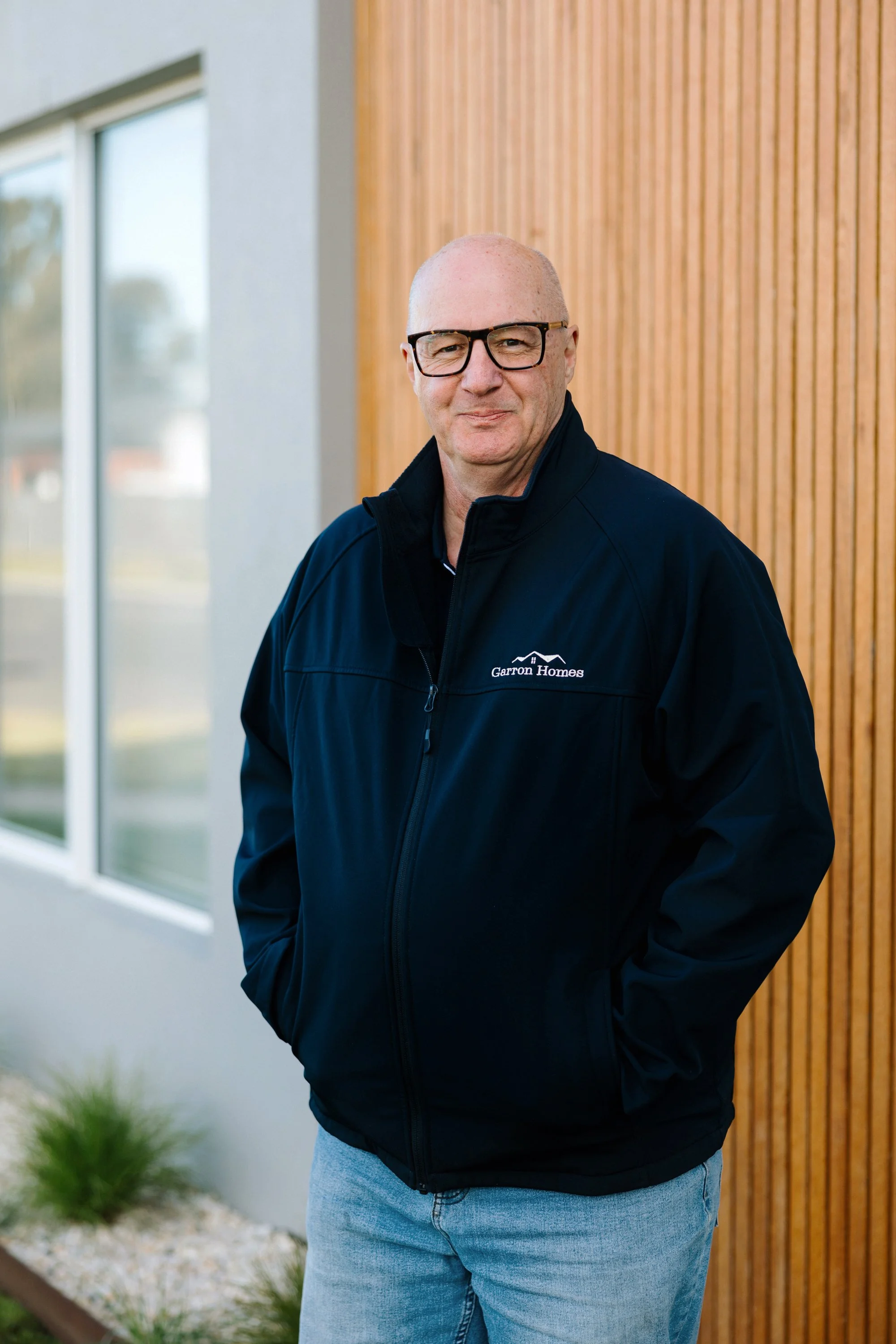 A bald man wearing glasses and a black jacket with 'Garron Homes' logo, standing outdoors near a modern building with wooden paneling and large windows.