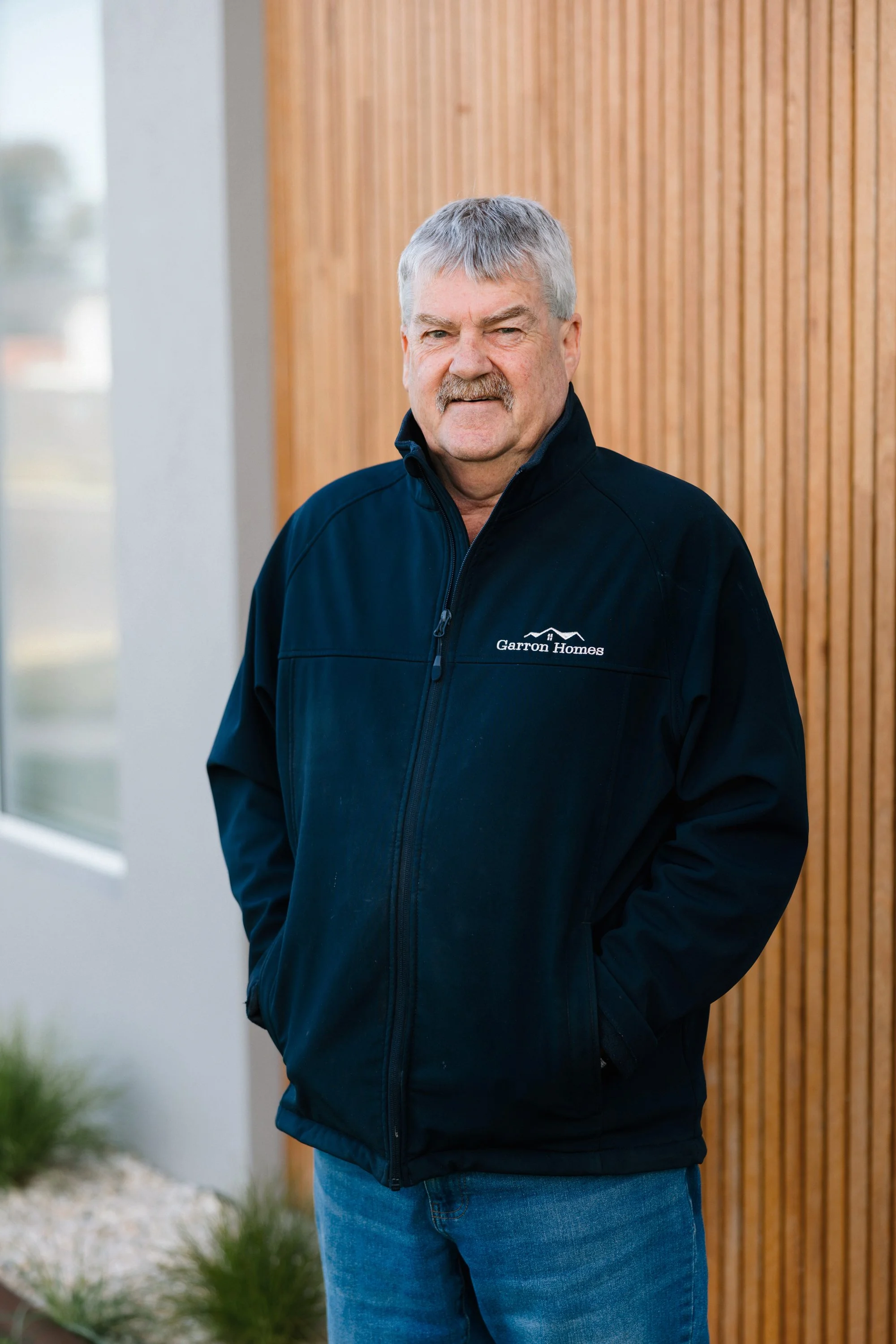 A man with gray hair and a mustache wearing a black Garro Homes jacket and blue jeans, standing outdoors against a wooden wall with vertical slats.