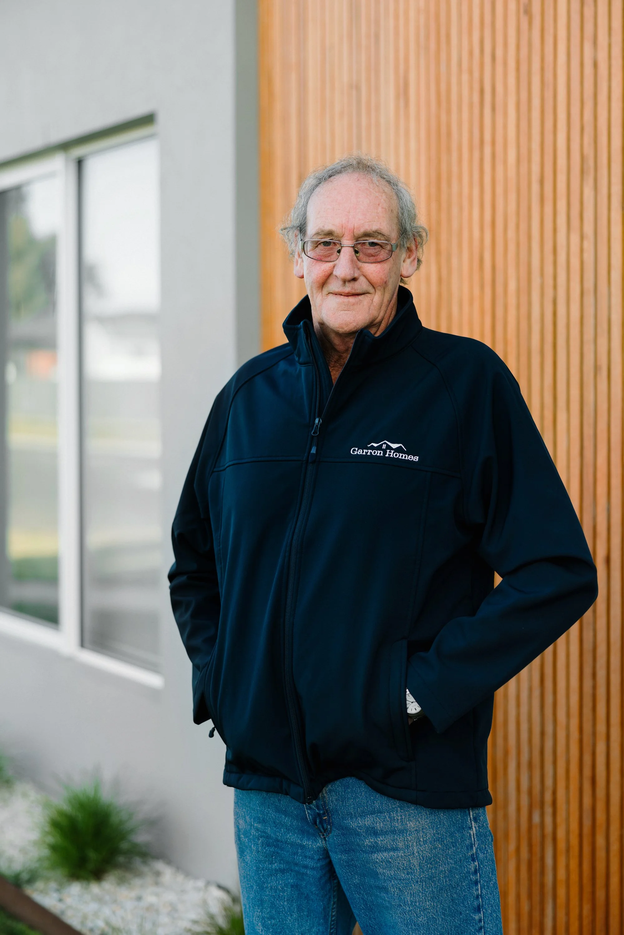 Man wearing glasses and a black jacket with 'Garron Homes' logo, standing outdoors with a wooden wall and window in the background.