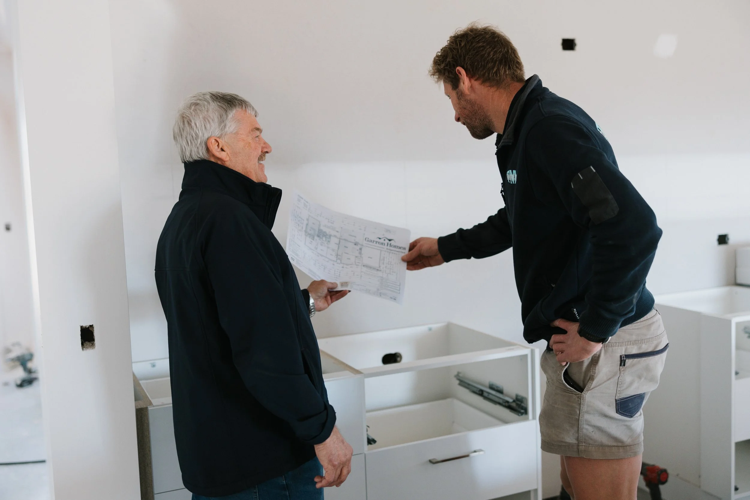 Two men, one older with gray hair, and one younger with brown hair, are inside a partially constructed kitchen. They are discussing a paper blueprint or plan. The kitchen cabinets are not yet installed, and there are exposed wall outlets and holes in the wall.