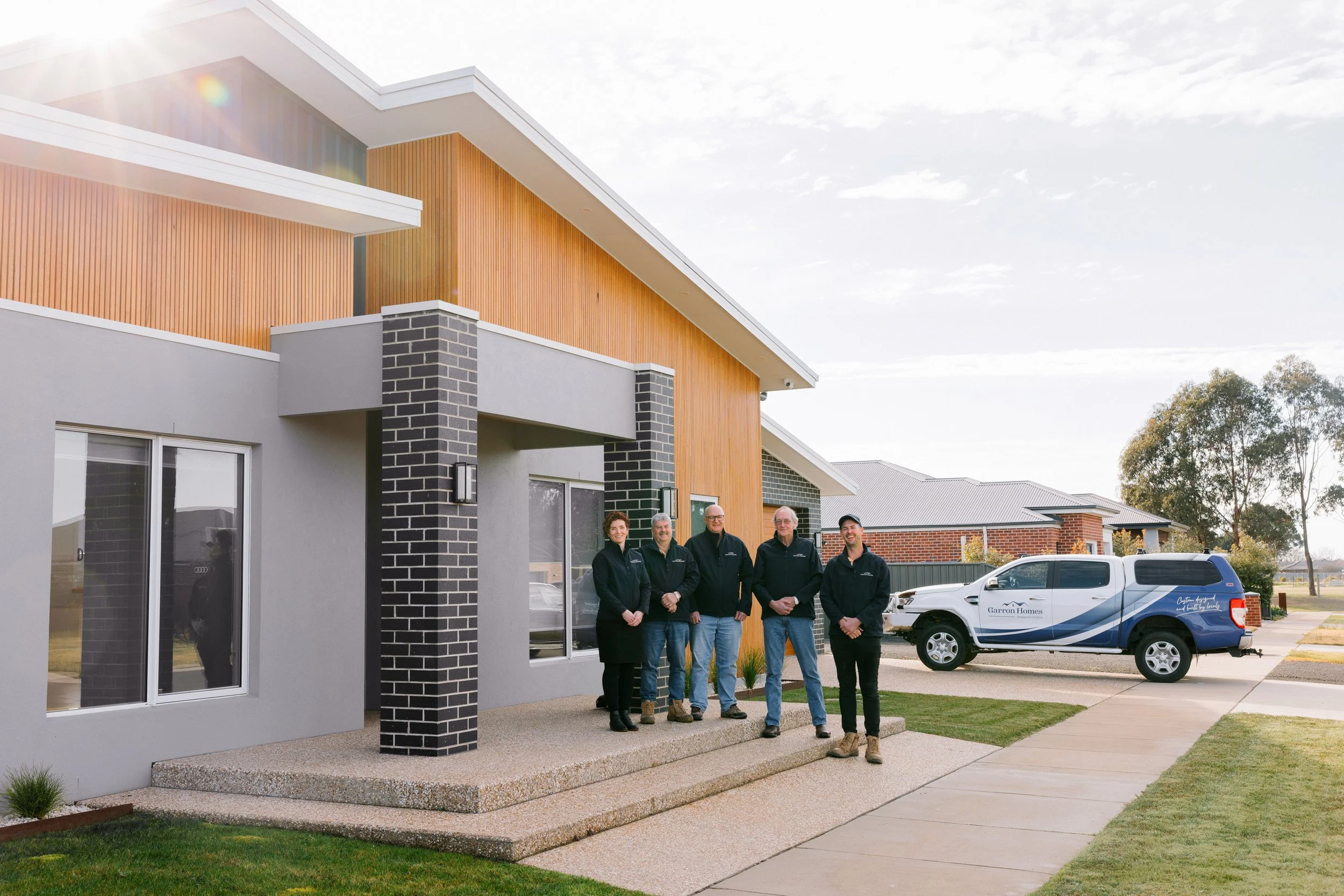 Group of five people standing in front of a modern residential building with a wooden and brick exterior, smiling and posing for the photo.