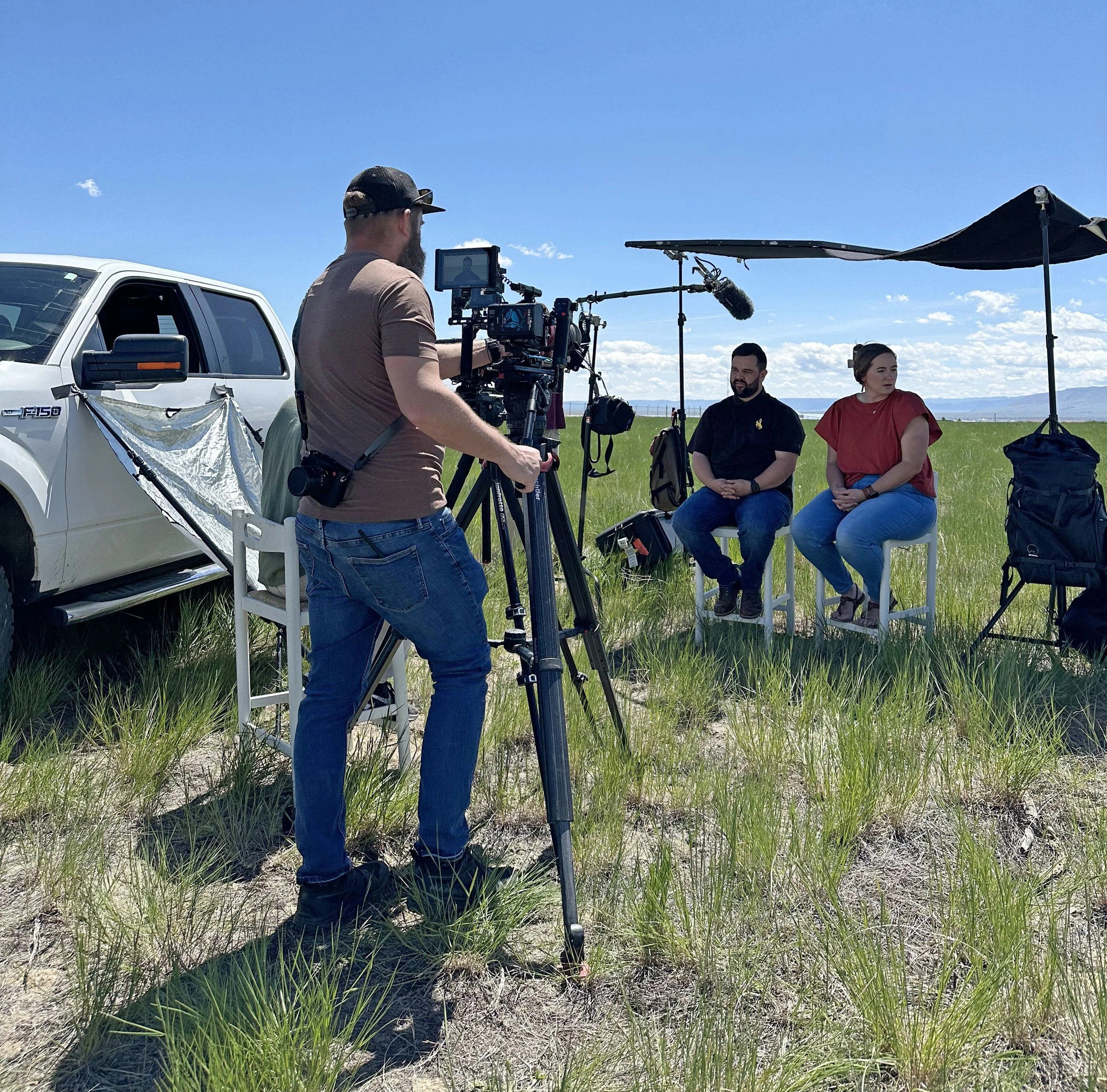 Filming crew setting up outdoor interview scene in a grassy field on a sunny day, with two people seated and a camera operator filming.