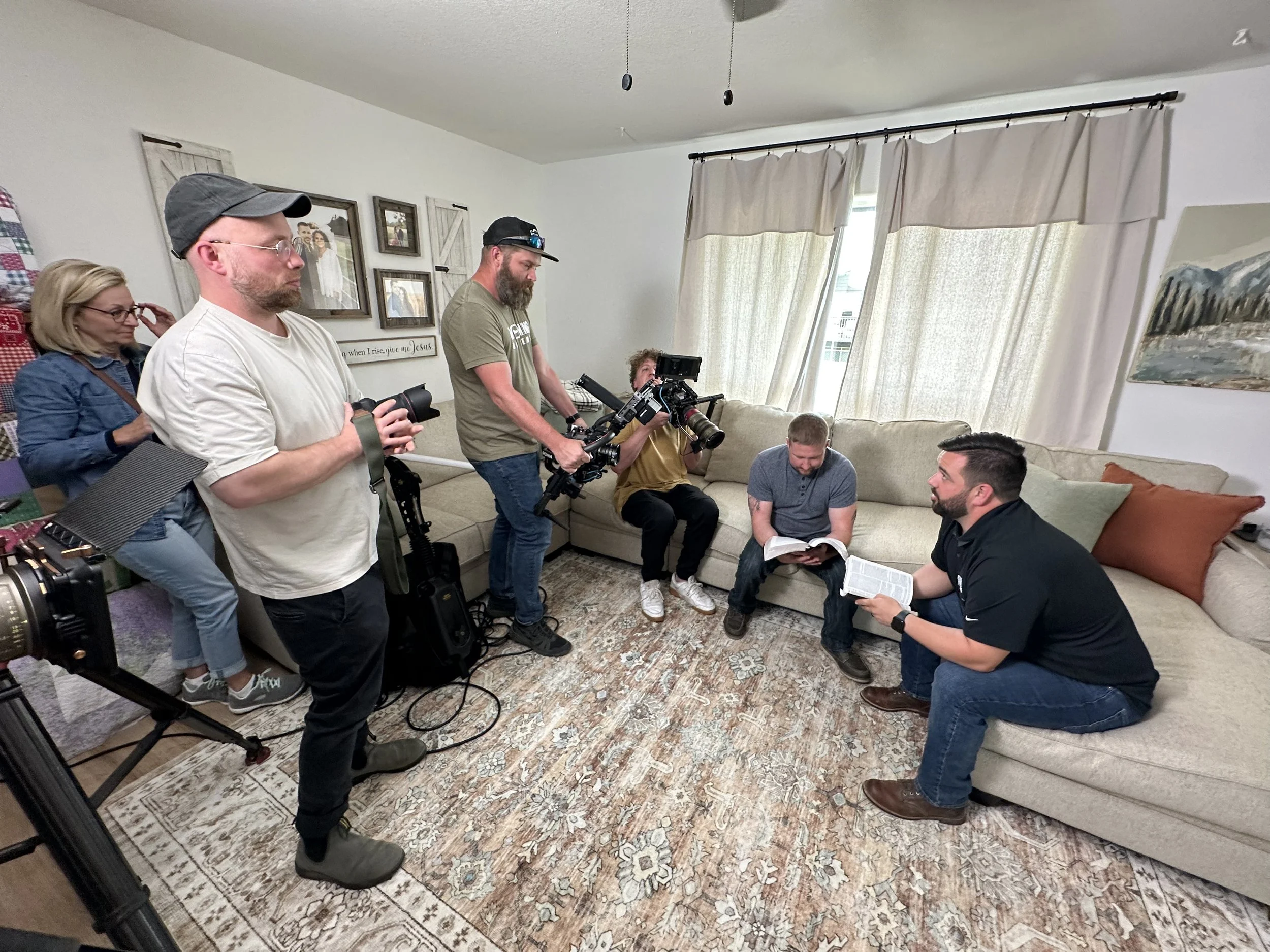 A group of six people in a living room filming or photographing a man sitting on a beige couch reading a book. Three are standing, with one holding a camera on a stabilizer, and two are sitting on the floor or couch with cameras. The room has a patterned rug, large windows with curtains, and wall art.