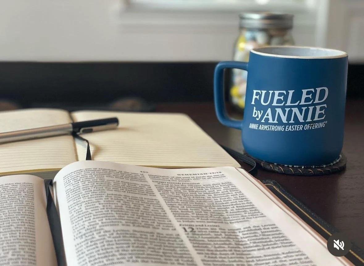 Open book and notebook with pen on a desk, blue mug with white text reading 'FUELED by ANNIE' and 'ANNIE ARMSTRONG EASTER OFFERING,' and a jar of colorful candies in background on a window sill.