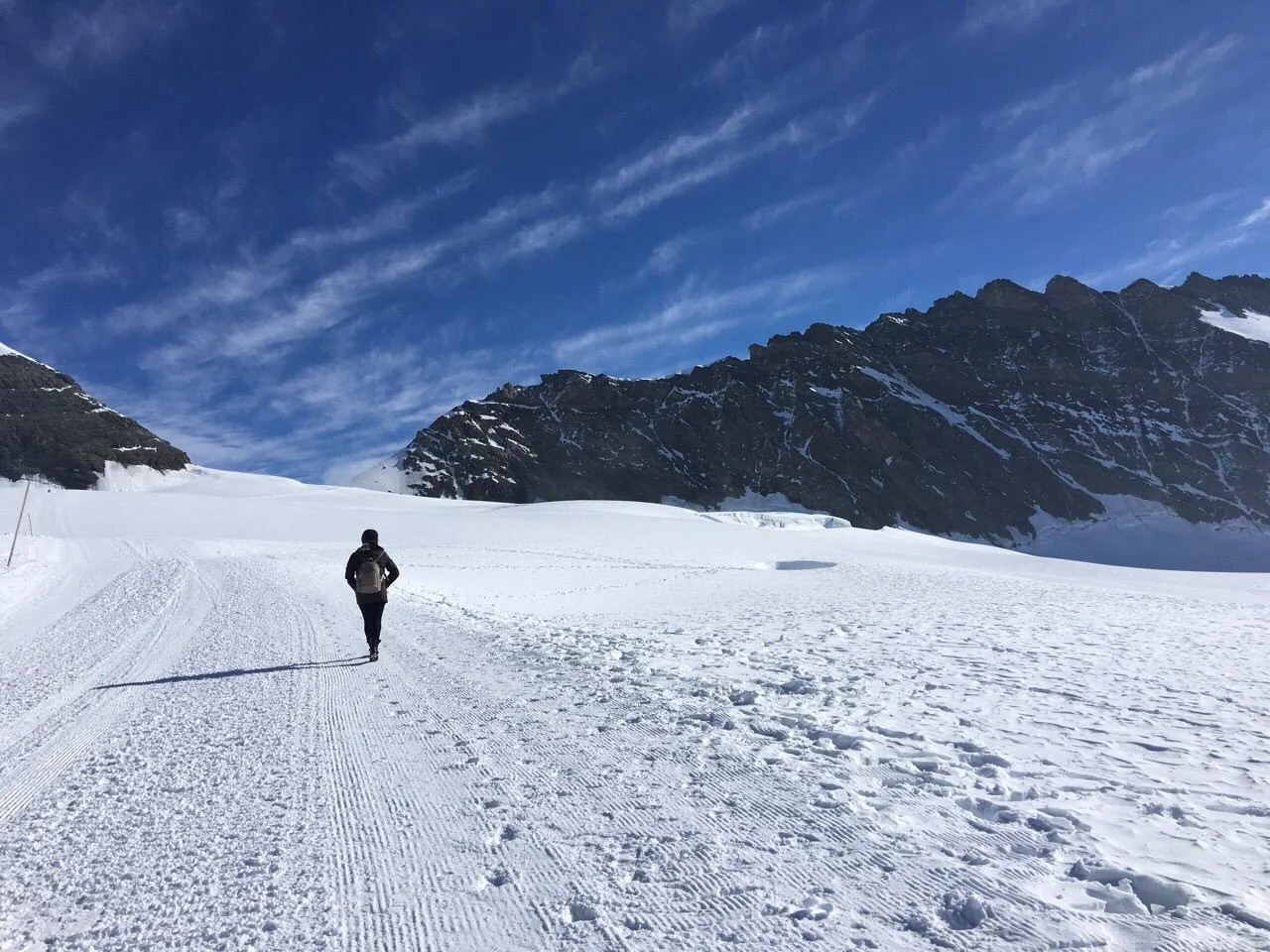 Person walking on snowy trail toward mountain range under a blue sky with wispy clouds.