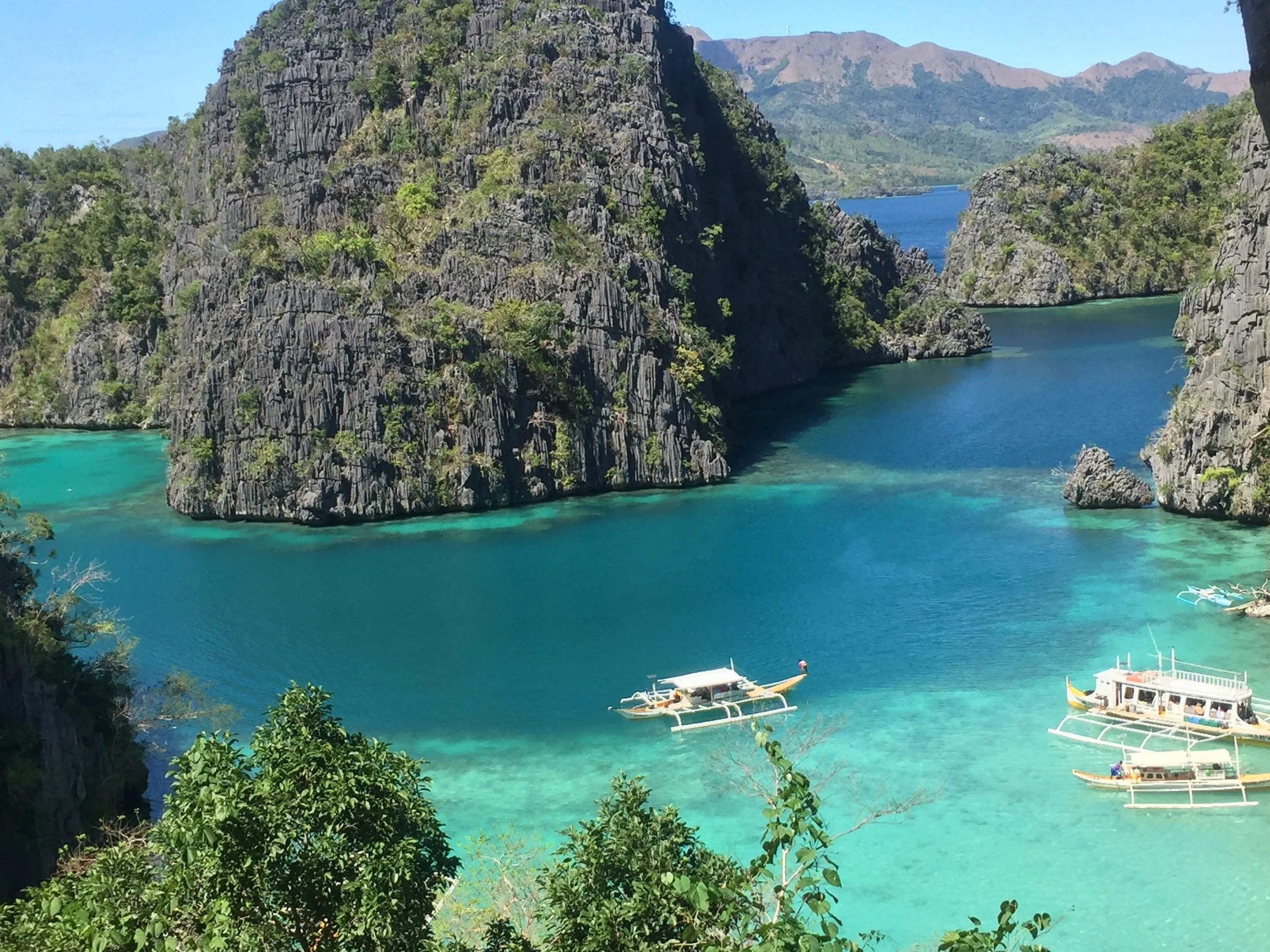Tropical bay with turquoise water, surrounded by tall, steep rocky cliffs covered with green vegetation, and several boats floating on the water.