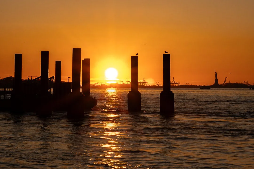 Sunset over the water with silhouetted posts and the Statue of Liberty in the background.