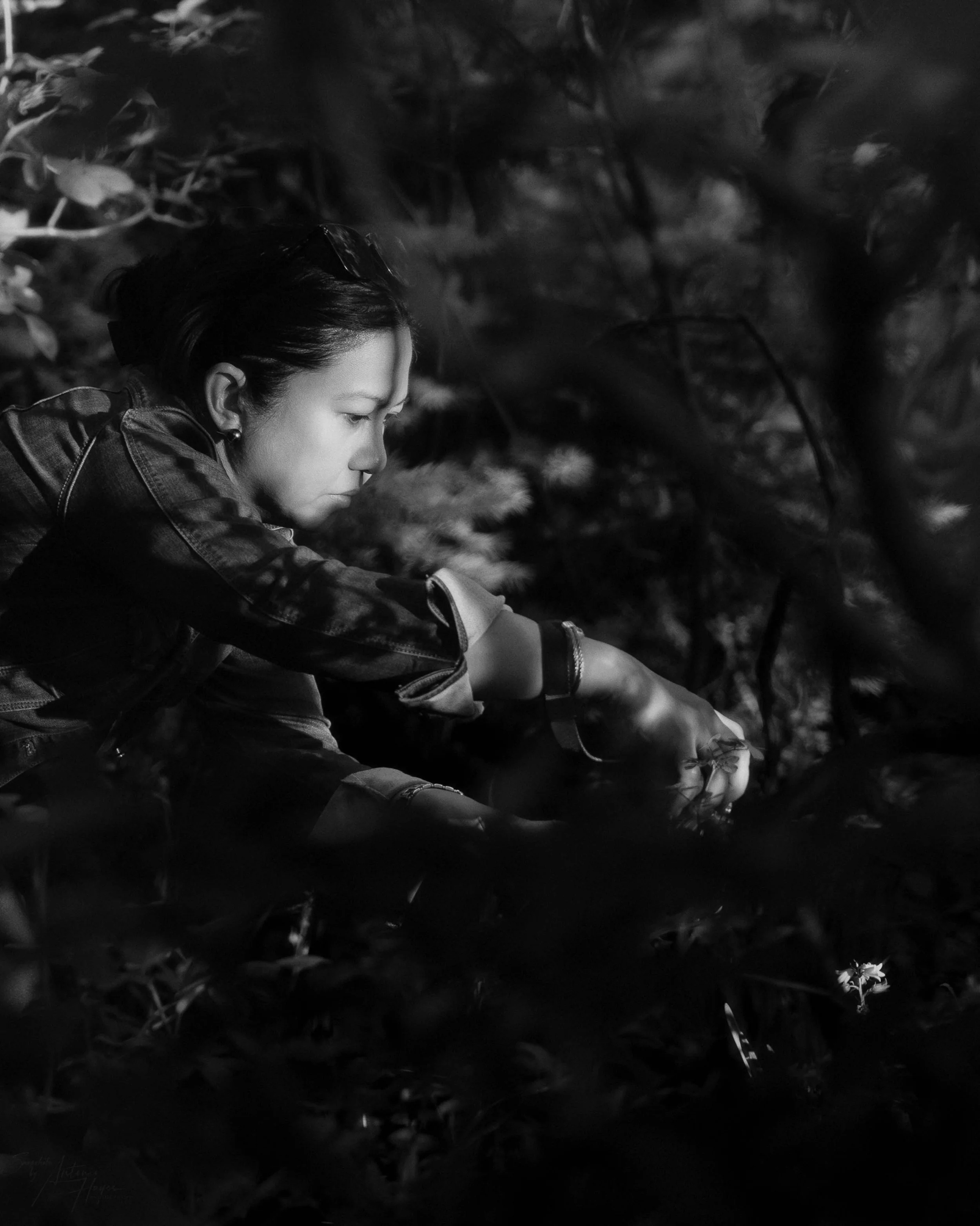 A woman with dark, shoulder-length hair and earrings, dressed in a denim jacket, crouches down outdoors among trees and plants at night, reaching for something on the ground.