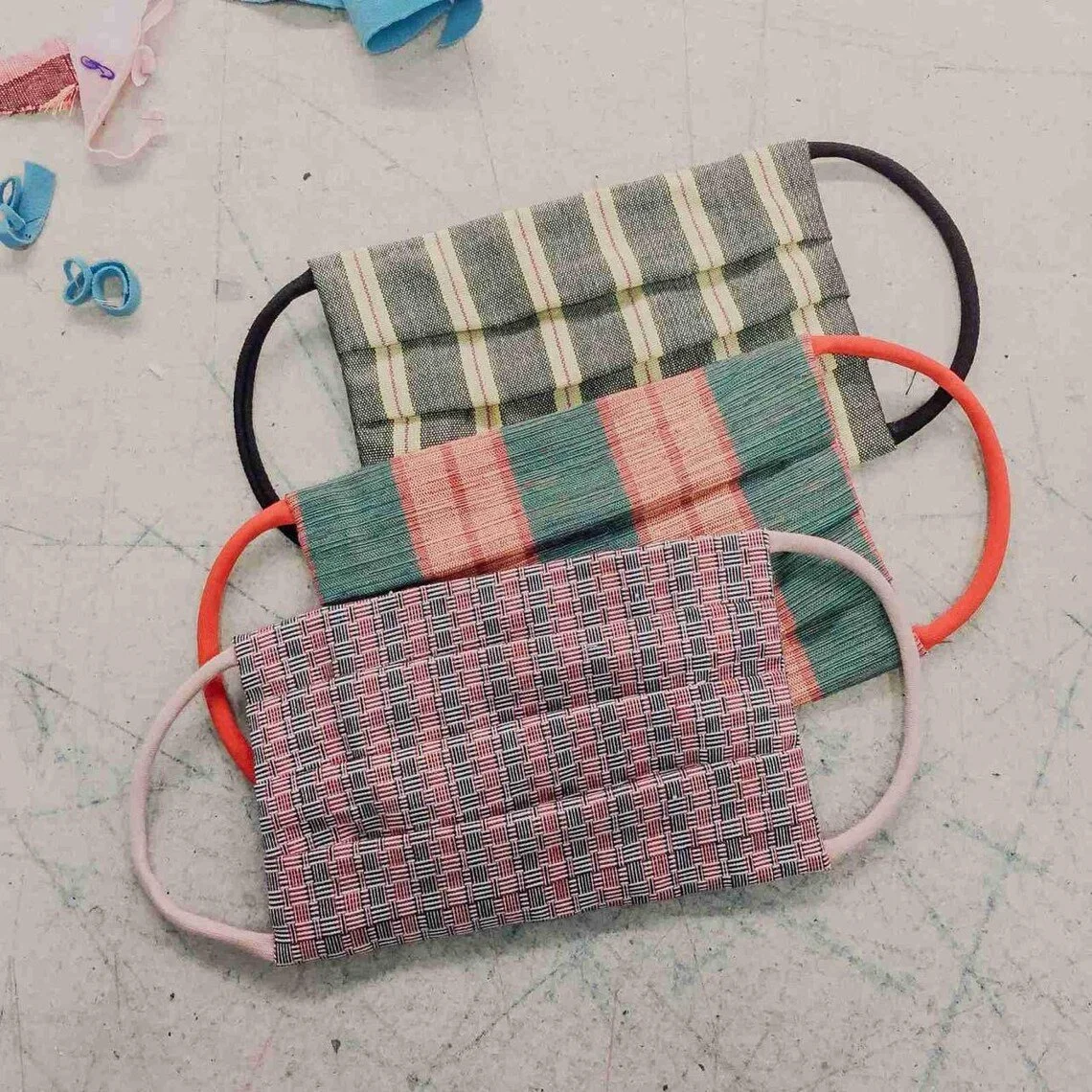Three fabric face masks with elastic ear loops laid out on a light-colored flooring. The top mask has a green and beige striped pattern, the middle mask has a red, green, and beige plaid pattern, and the bottom mask has a red, black, and white checkered pattern. There are small fabric scraps and rubber bands scattered around.