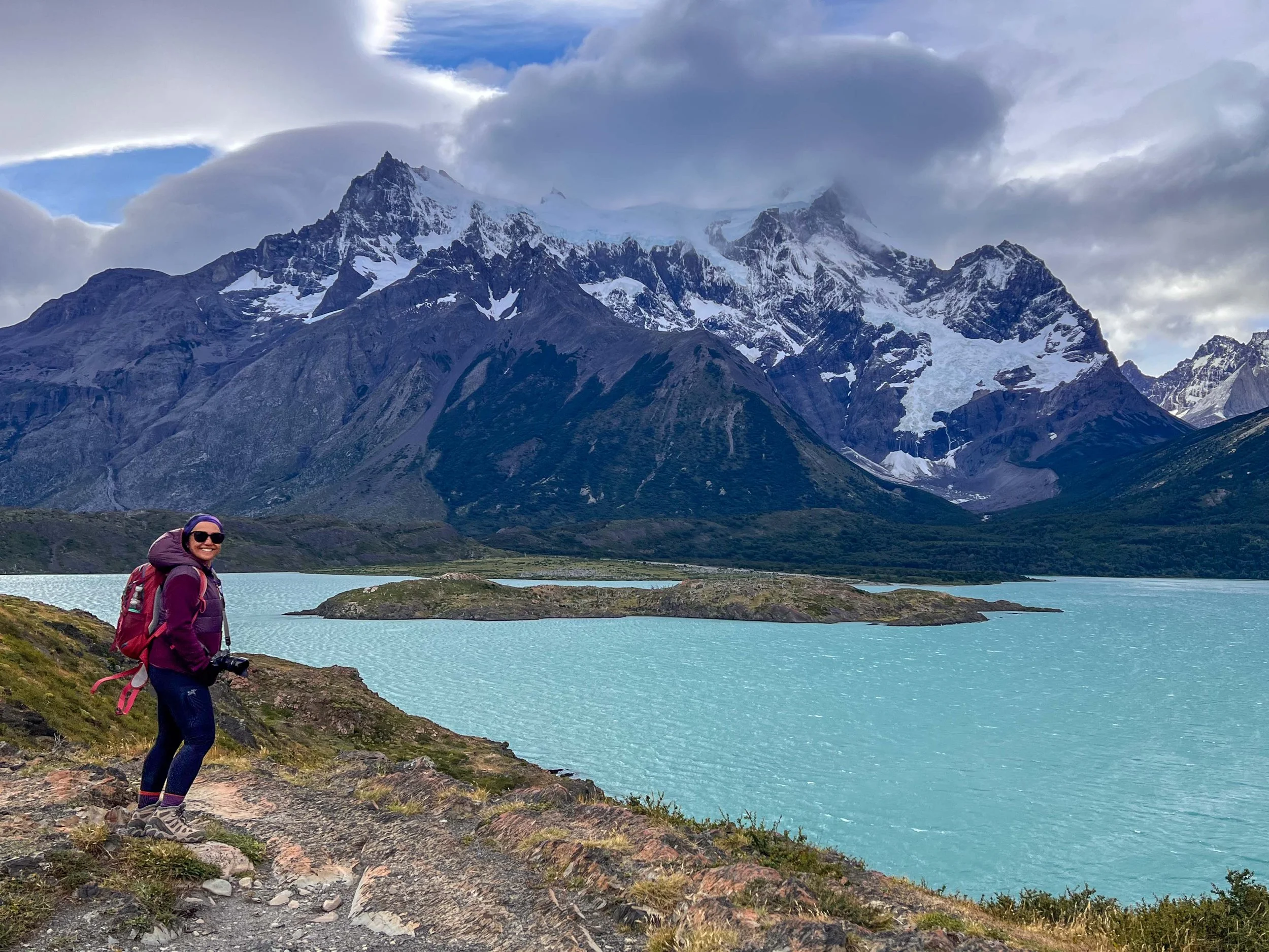A woman hiking near a lake with snow-capped mountains in the background.
