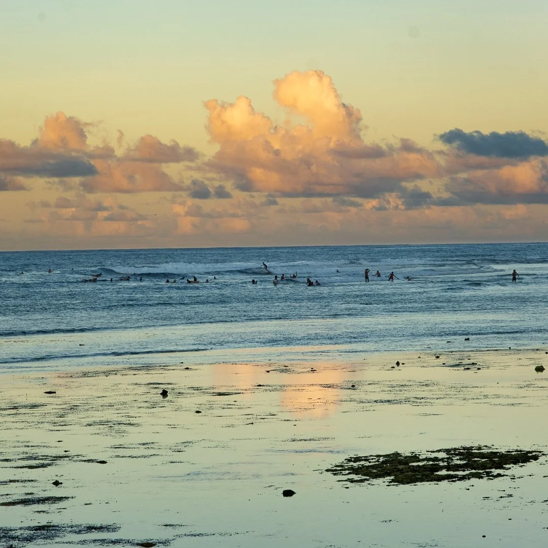 Beach scene with people surfing and swimming in the ocean during sunset, with clouds reflecting in the wet sand.