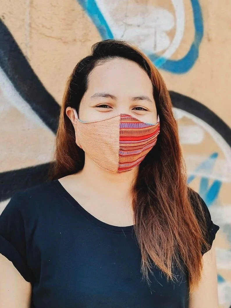 A smiling woman with long brown hair wearing a colorful striped face mask and a black shirt, standing in front of a graffiti wall.