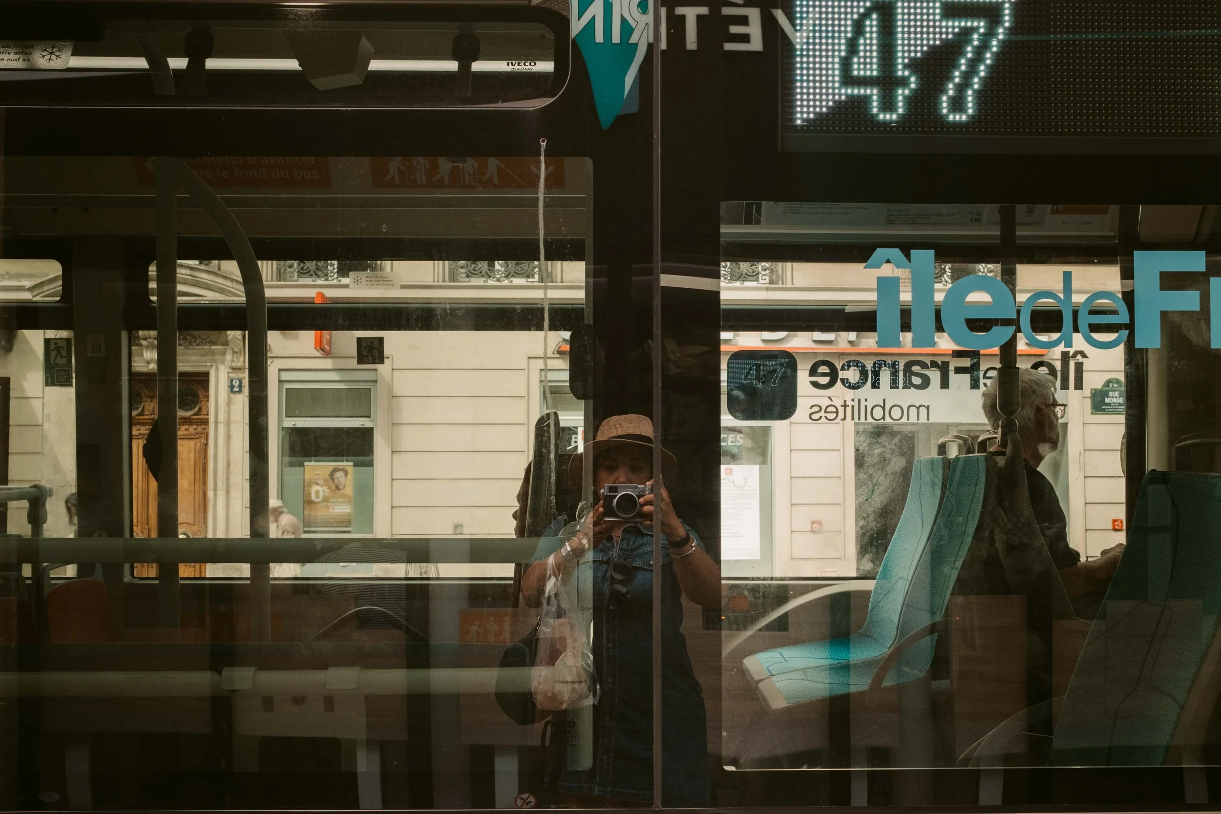 Reflection of a woman taking a photo with a camera inside a bus, with city buildings visible outside through the bus window.