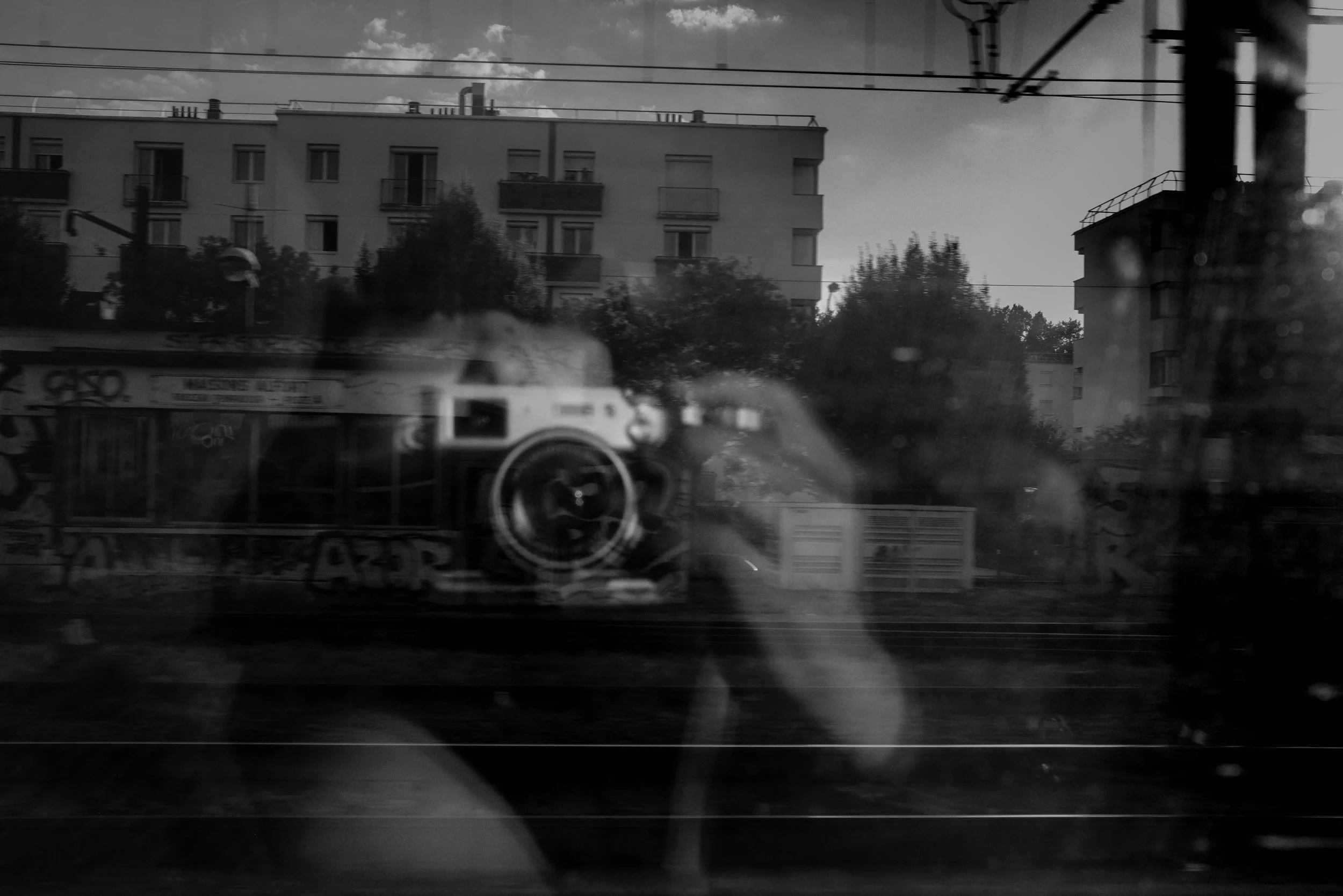 Black and white photograph of a cityscape reflected in glass, showing buildings, electrical wires, and a person taking a photo with a camera, with a graffiti-covered train or train car in the foreground.