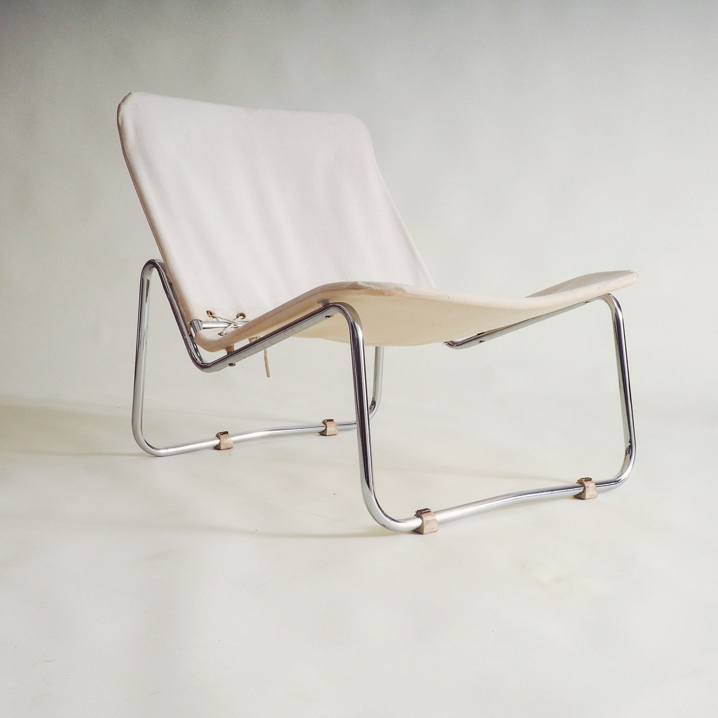 70s tubular lounge chair

Minimalistic vintage lounge chair featuring a polished chrome tubular frame and a white fabric sling seat and backrest. The white fabric is attached to the chrome frame with a lacing system, visible on the sides where eyelet