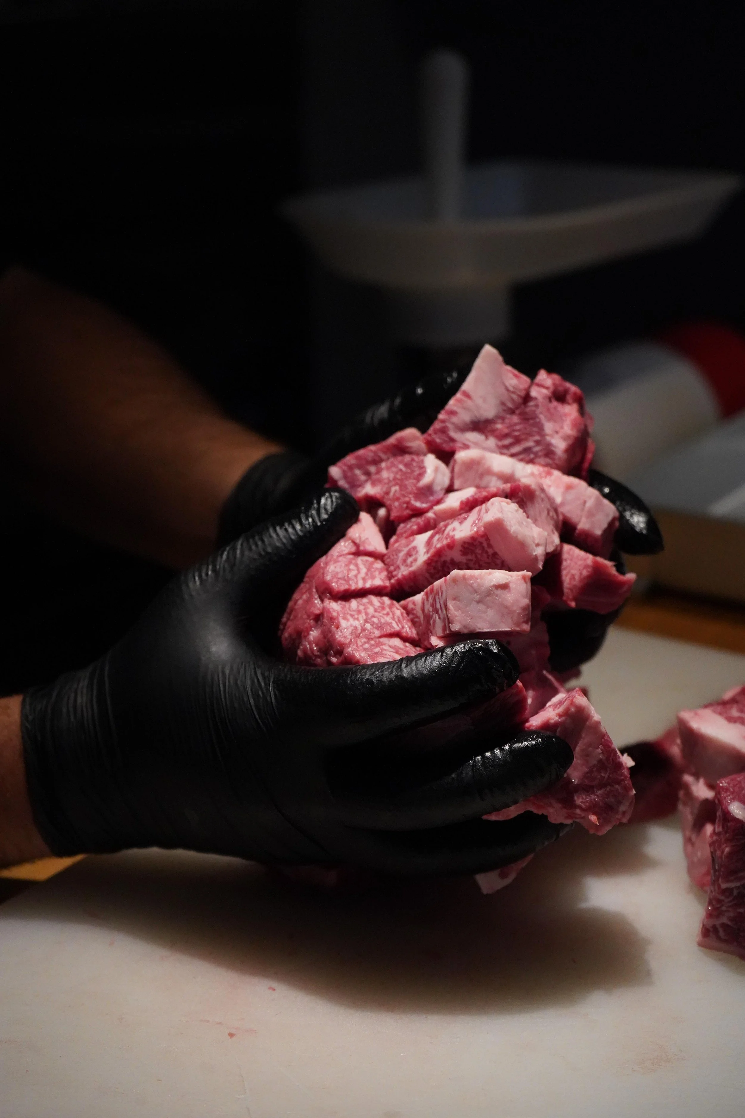 Person wearing black gloves handling raw, cubed beef meat on a white cutting board in a kitchen.