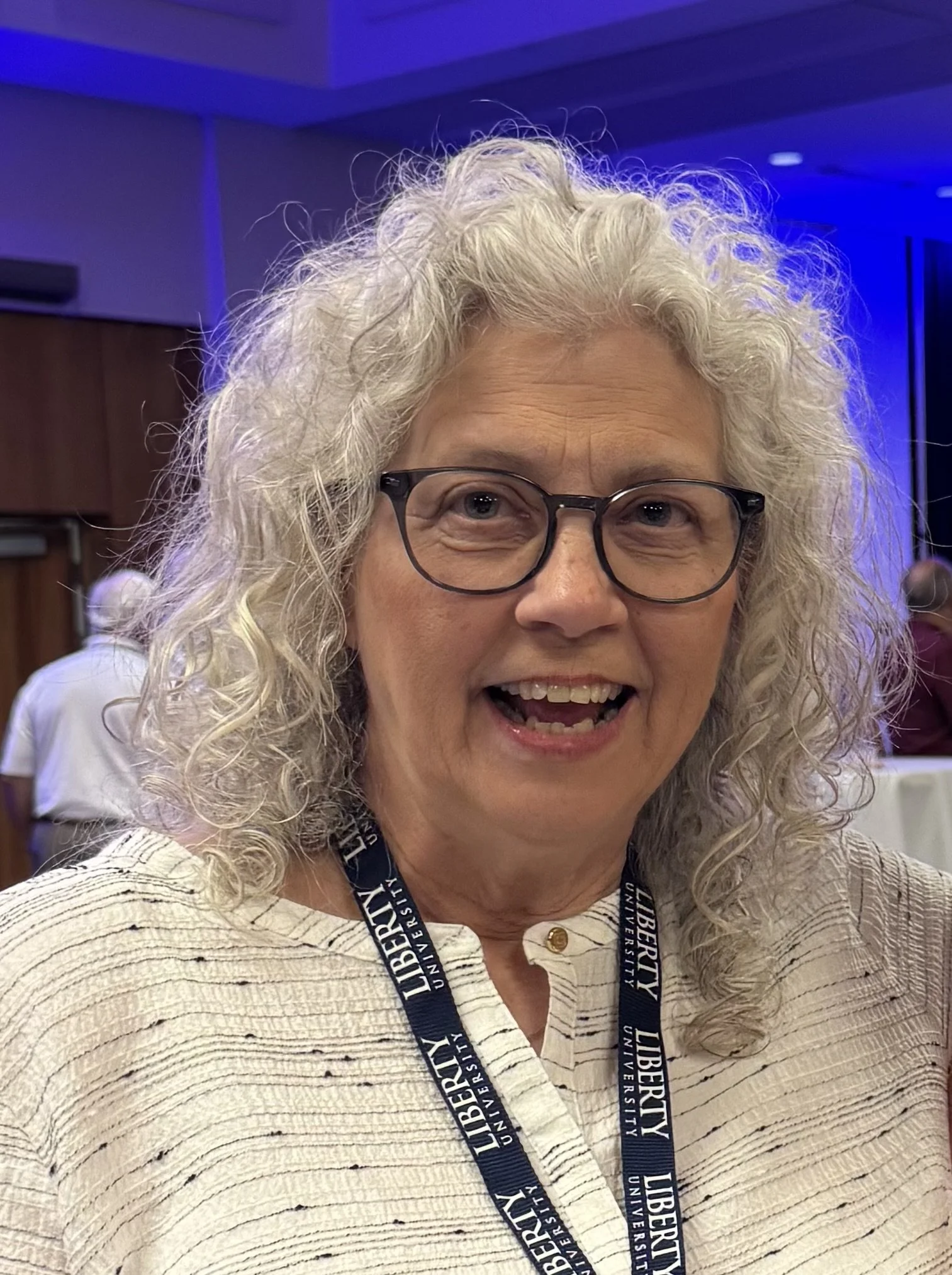 A smiling woman with curly gray hair and blue eyes, wearing a white top.