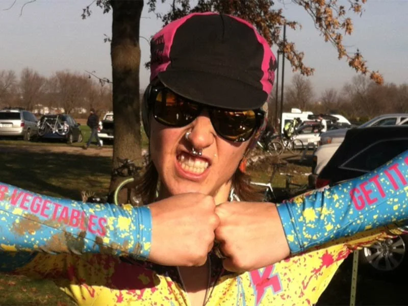 Person wearing sunglasses, a black and pink cap, and a brightly colored, spattered long-sleeve shirt, with arms crossed in a confident pose. Background shows parked cars, bicycles, and trees.