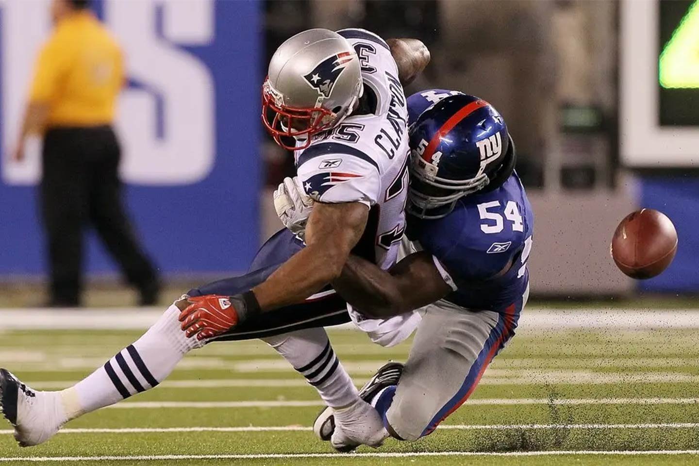 A football player from the New York Giants falls while tackling a player from the New England Patriots, with the fumbled football flying in the air.
