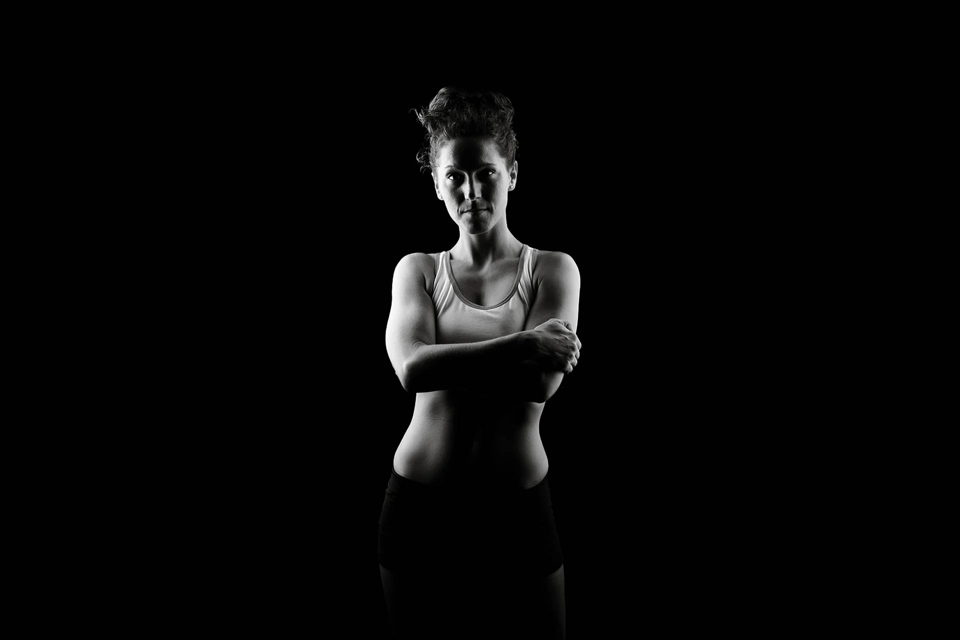 Black and white photo of a woman standing with her arms crossed, in a sleeveless top, against a dark background.