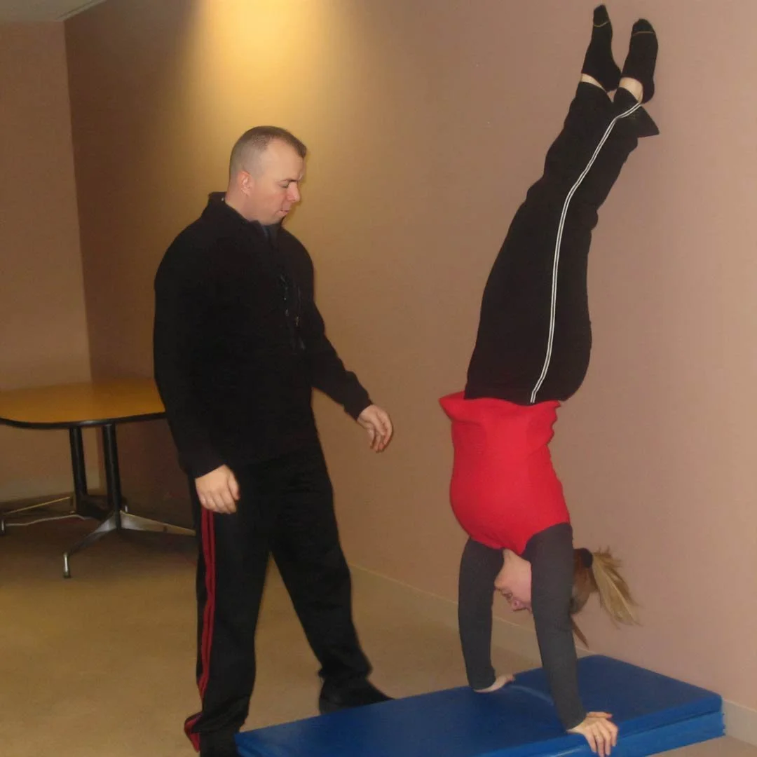 A woman doing a handstand on a blue mat with her legs against a wall, while COACH RHINO watches beside her.