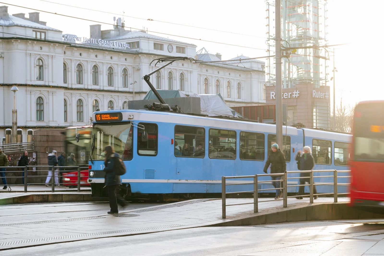 The Oslo Central Station area pulses with the city's energy, where the iconic blue tram carves a path through the frame. Crisp Nordic light illuminates the commuter flow against the classic facade of the Grand Central Hotel, capturing the core inters