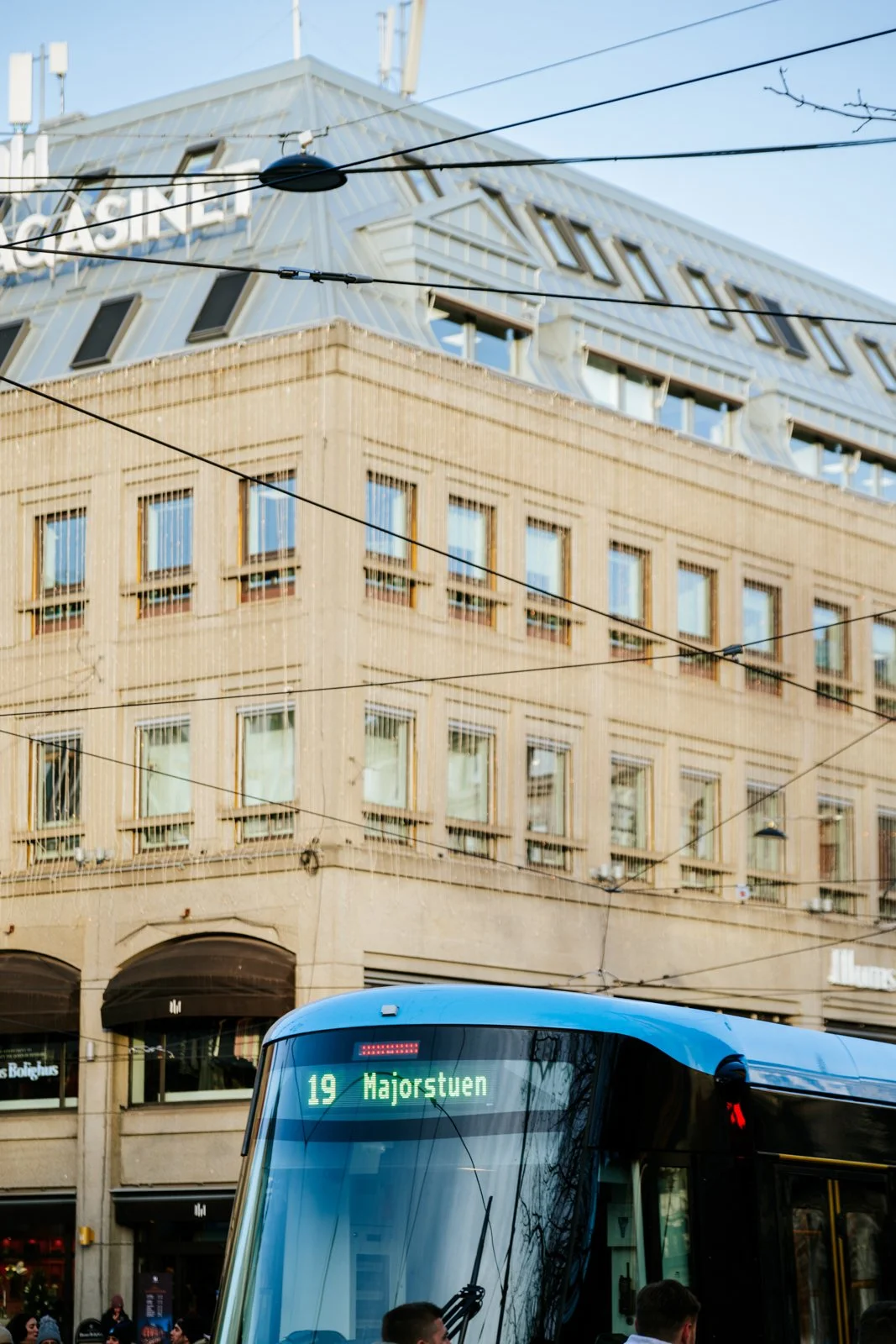 A modern blue tram on a city street with a digital display showing route 19 to Majorstuen, passing in front of a beige multi-story building with numerous windows and electric wires overhead.