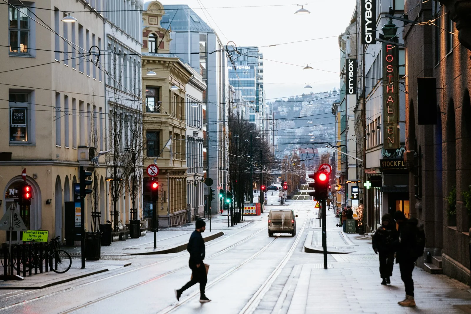 City street with buildings on both sides, some with neon signs. Several pedestrians cross the street, and a vehicle drives through an intersection with red traffic lights. Overhead wires are visible, and in the background, hills and more buildings ca