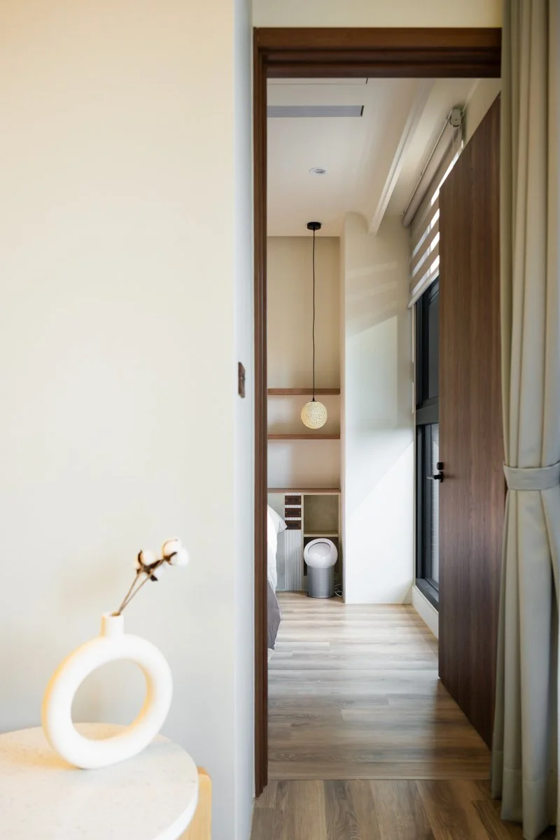 View of a hotel room hallway with wooden floors and a sliding door, revealing a bed, hanging pendant light, and an air purifier inside the room. 中性色調的宜蘭住宅，以質感提升空間層次。