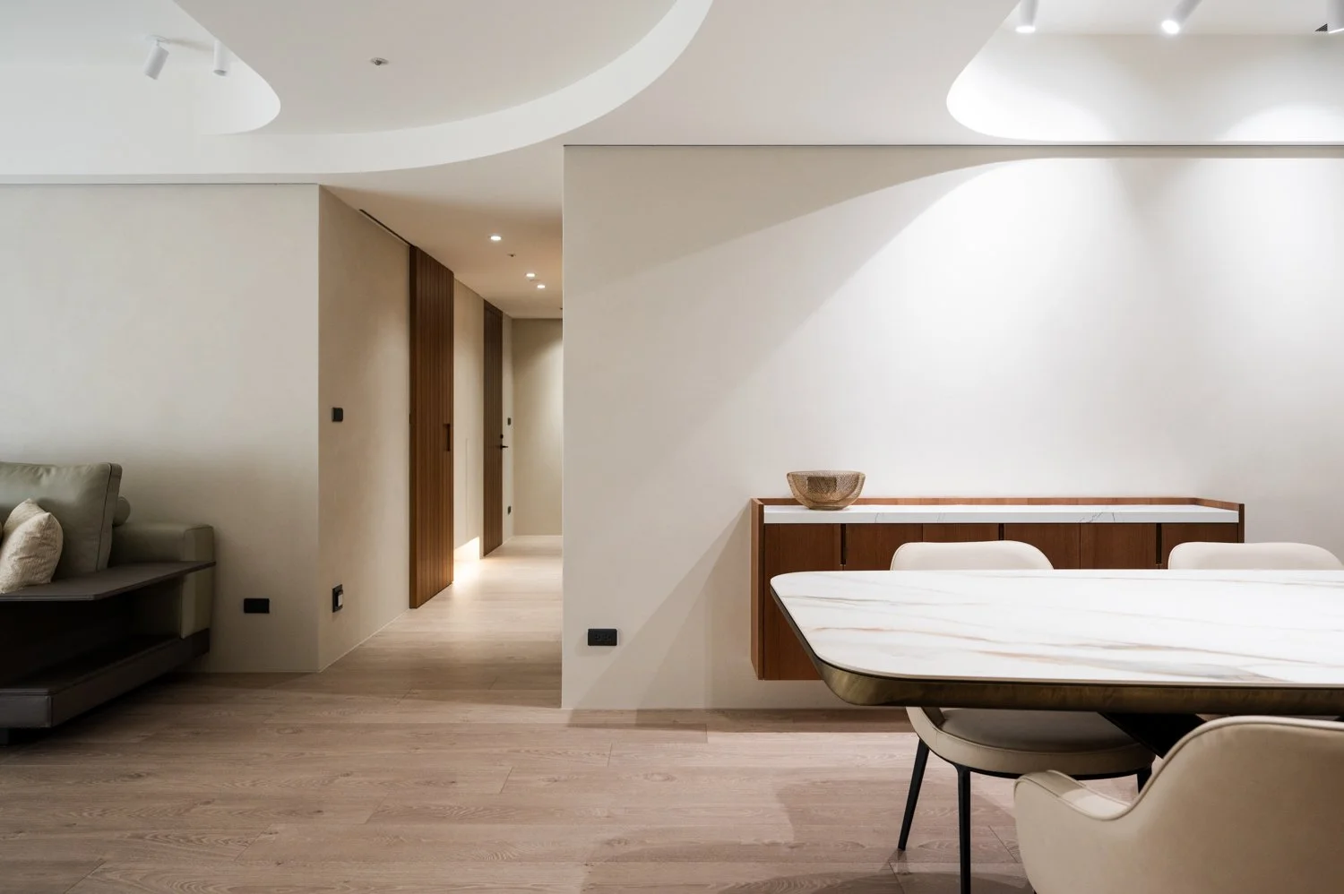 Modern living room with light wood flooring, beige walls, and minimalist furniture including a marble top dining table, cream chairs, a sideboard with a decorative bowl, and a green sofa with cushions. Hallway with wooden sliding doors is visible in the background.