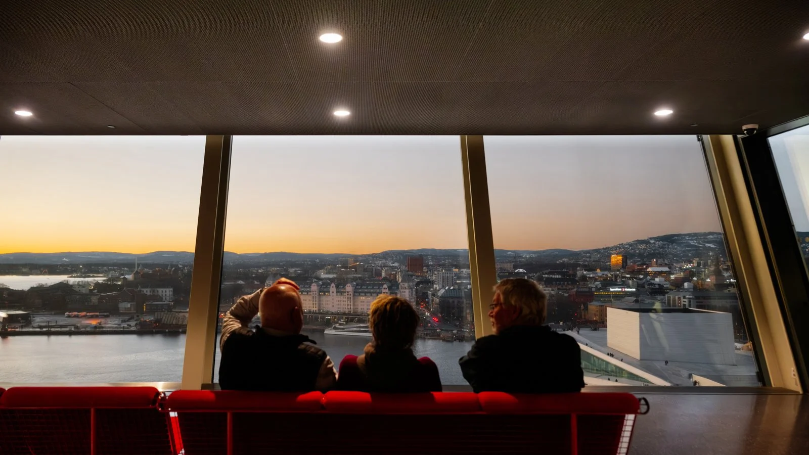 Three people sitting on a red bench in front of large windows overlooking a city skyline and body of water at sunset.