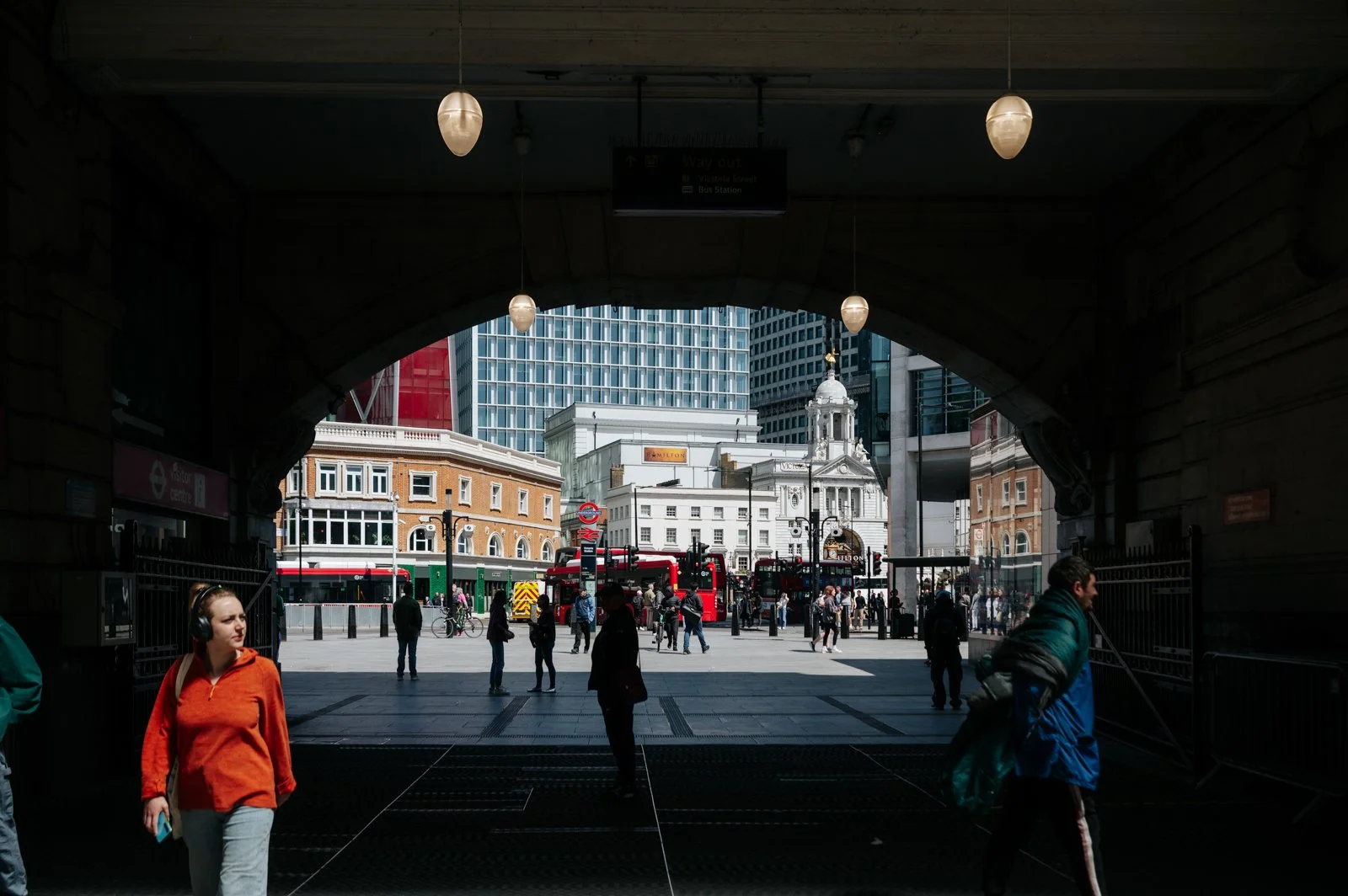 View of a busy city square in London, seen from under a tunnel. People walk across the square, with iconic red buses and historic buildings, including a church with a white facade and a clock tower, in the background.