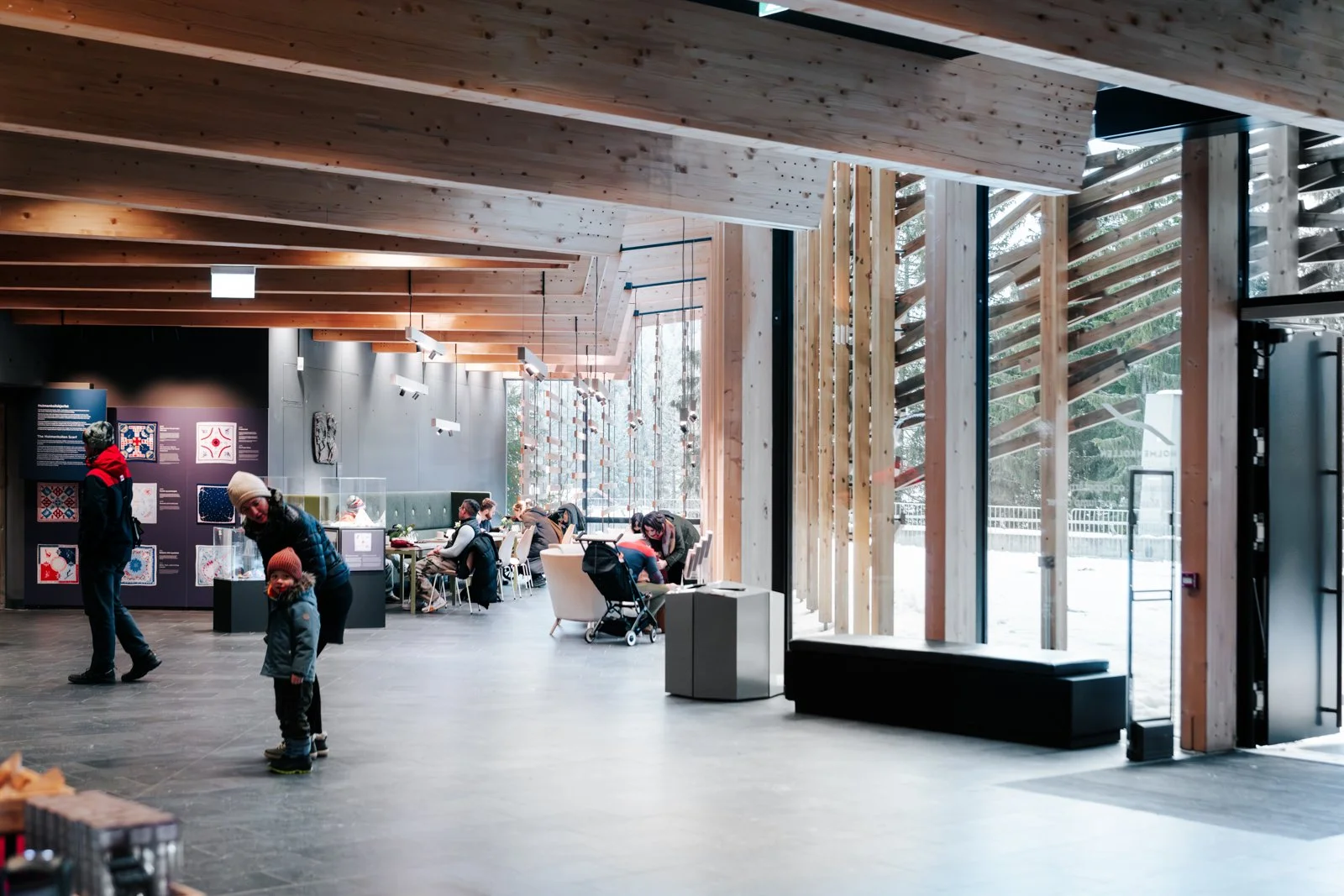 Interior of a modern building with wooden ceiling beams, large windows, and several people sitting and standing, including a woman with a child and others at tables, with artwork displayed on walls.