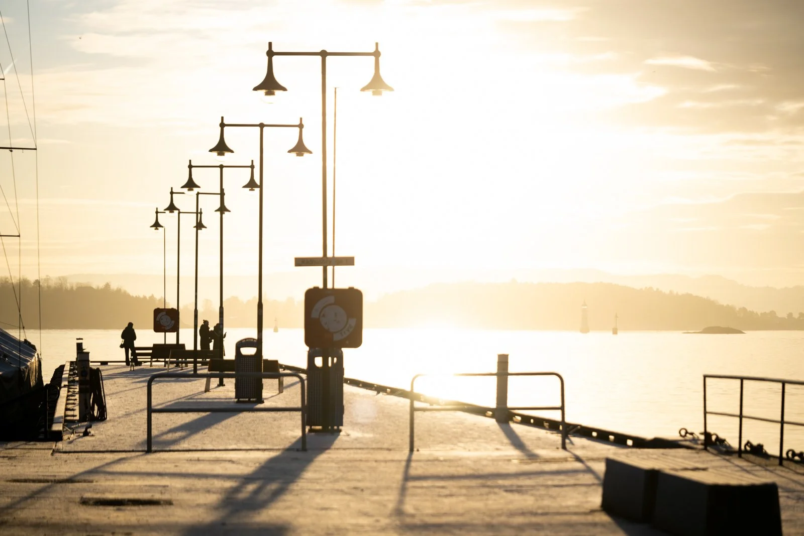 The Oslo Fjord at golden hour is captured in a stunning display of light and depth. The rhythmic row of pier lampposts creates a strong leading line and a dramatic silhouette against the blindingly bright horizon. The low sun bathes the scene in a wa