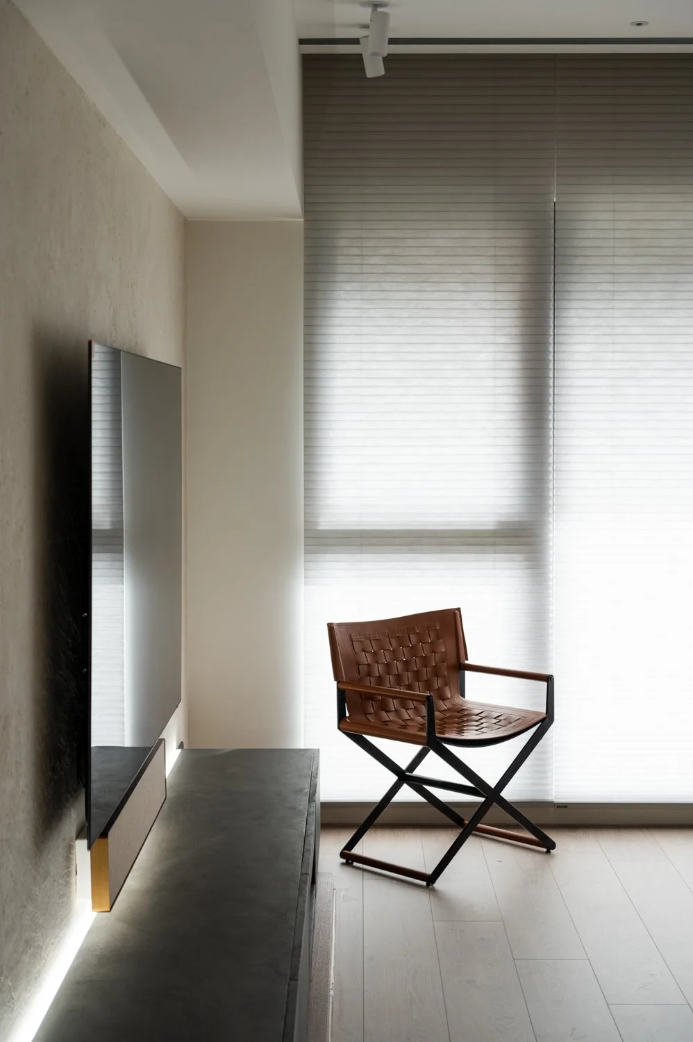 Minimalist living room with a flat-screen TV on a console, a brown woven leather chair, and large windows with white blinds letting in natural light.
