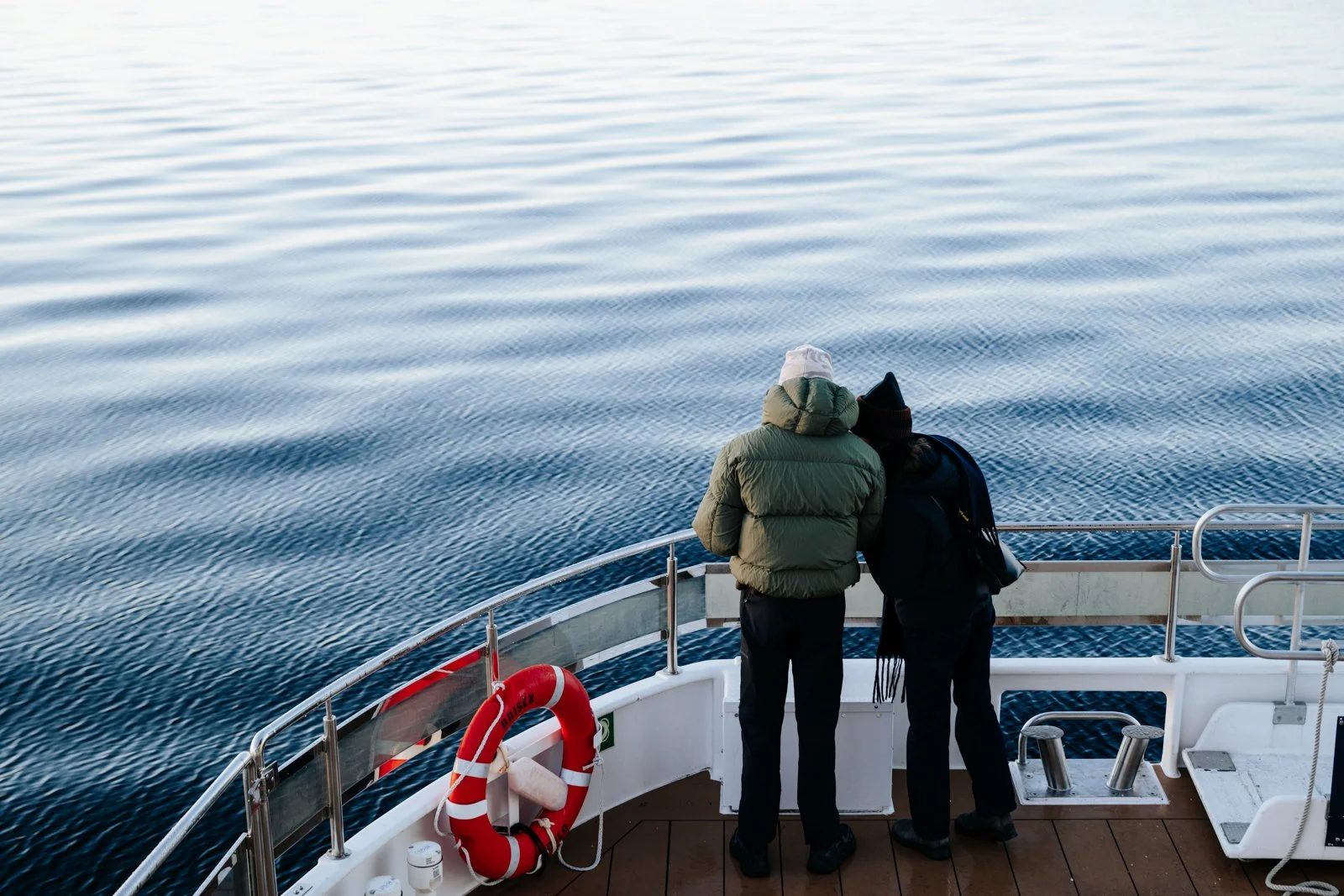 A view from the deck of a fjord cruise, capturing two figures absorbed in the vast, rhythmic water-scape. The clear blue surface is defined by intricate, crisp ripples, highlighting the boat's motion. The image conveys a sense of quiet companionship 