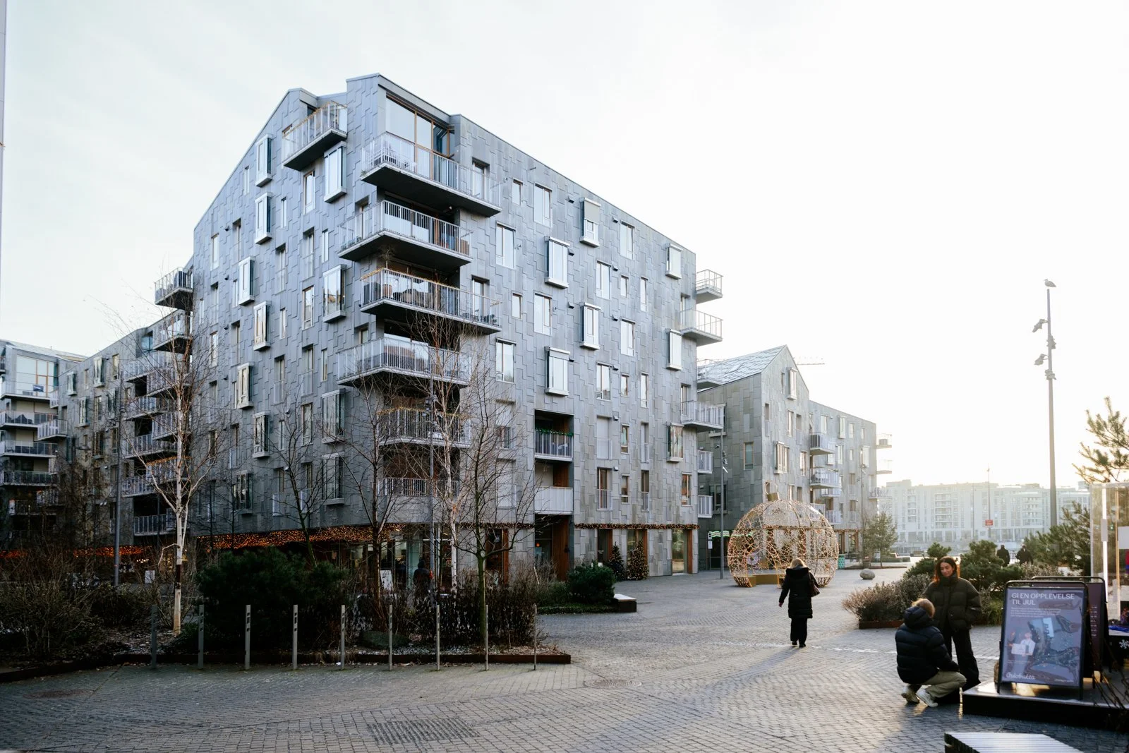 Modern residential apartment complex with multiple stories, balconies, and varied window sizes, set in an urban environment with a paved pedestrian area, trees, and decorative outdoor structures, with people walking and sitting nearby.