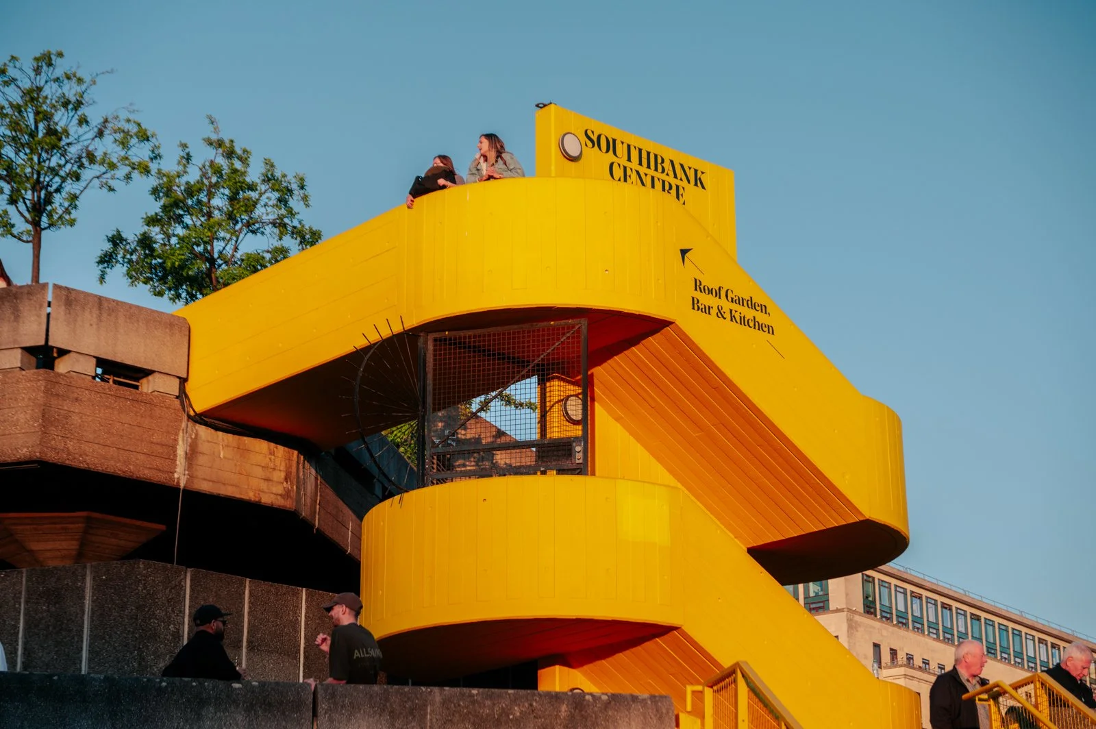 Yellow spiral staircase with people sitting on top, sign reading 'Southbank Centre' and directions to the rooftop garden, bar, and kitchen, against a clear blue sky.