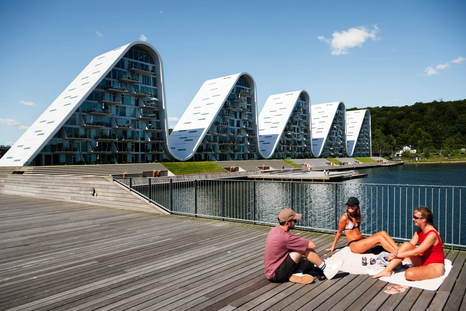 People enjoying the public boardwalk beneath The Wave's facade. Emphasizing the structure's integration with Vejle Fjord's leisure space.