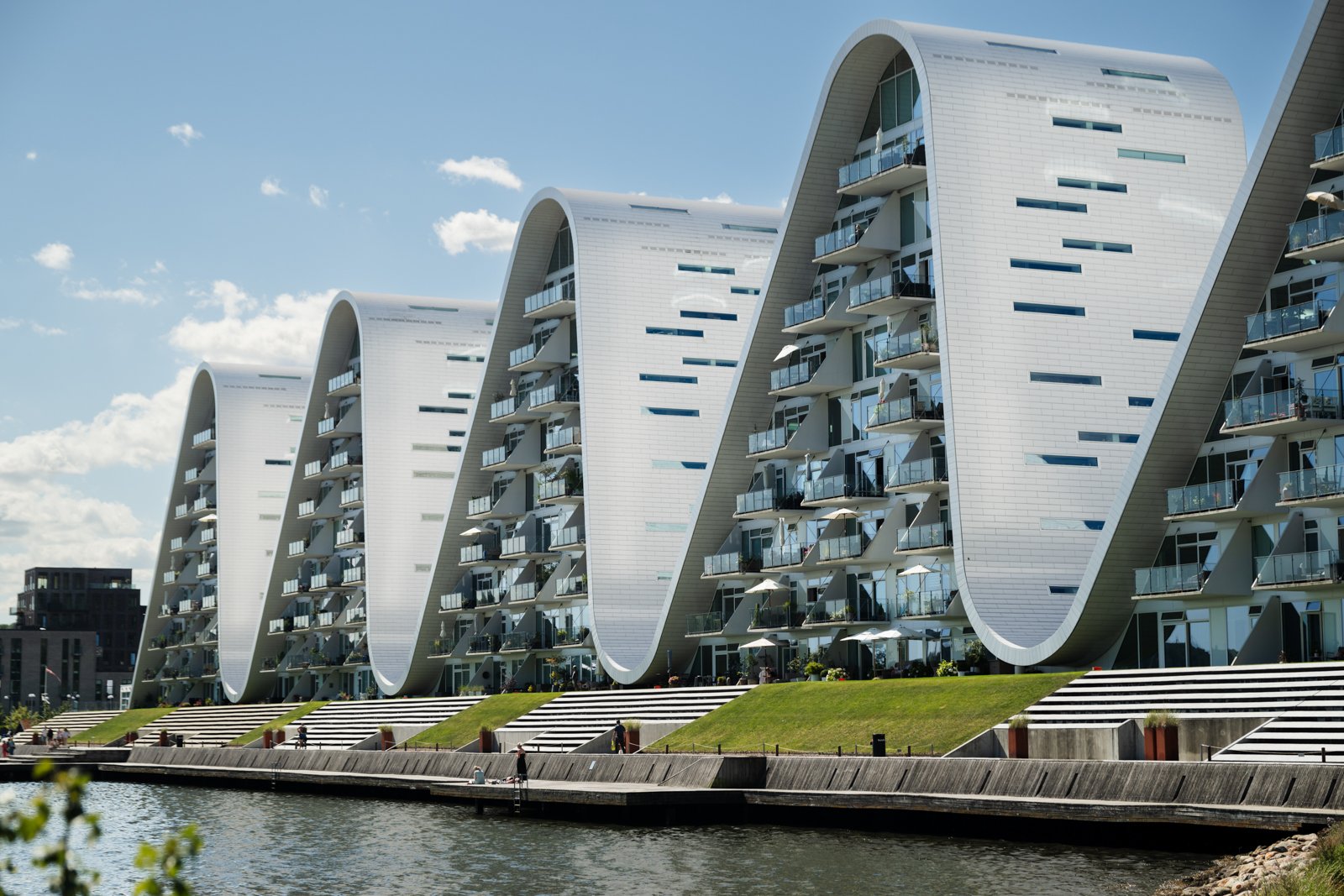 Architectural rhythm of the five wave segments reflecting on the calm fjord water, viewed from the natural park edge.