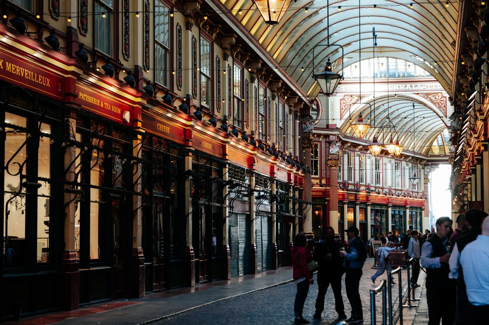 People walking inside a shopping arcade with ornate architectural details, glass ceiling, chandeliers, and storefronts.