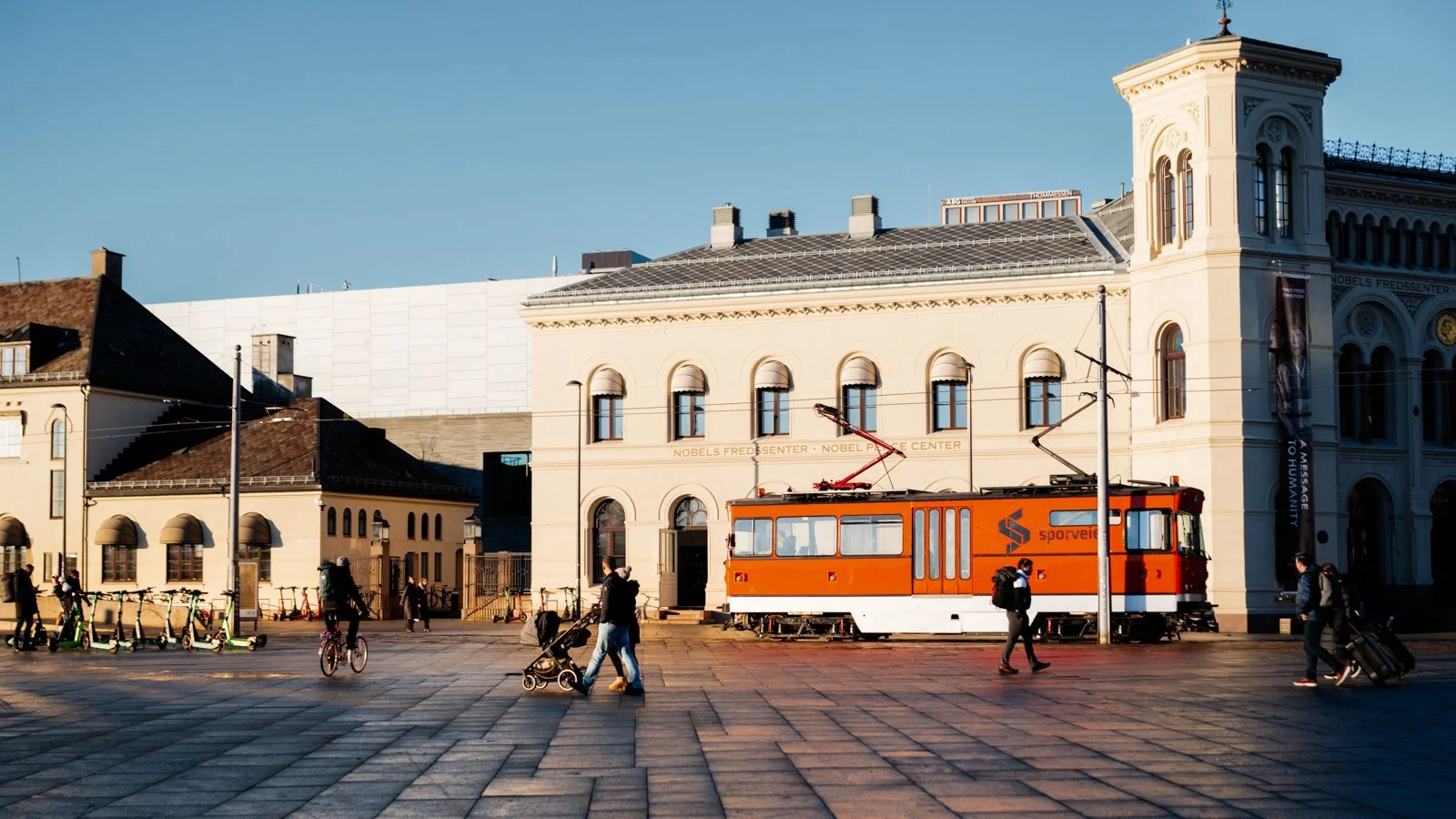 People walking on a city square with a vintage orange tram, surrounded by buildings, some with arched windows, and a clear blue sky.