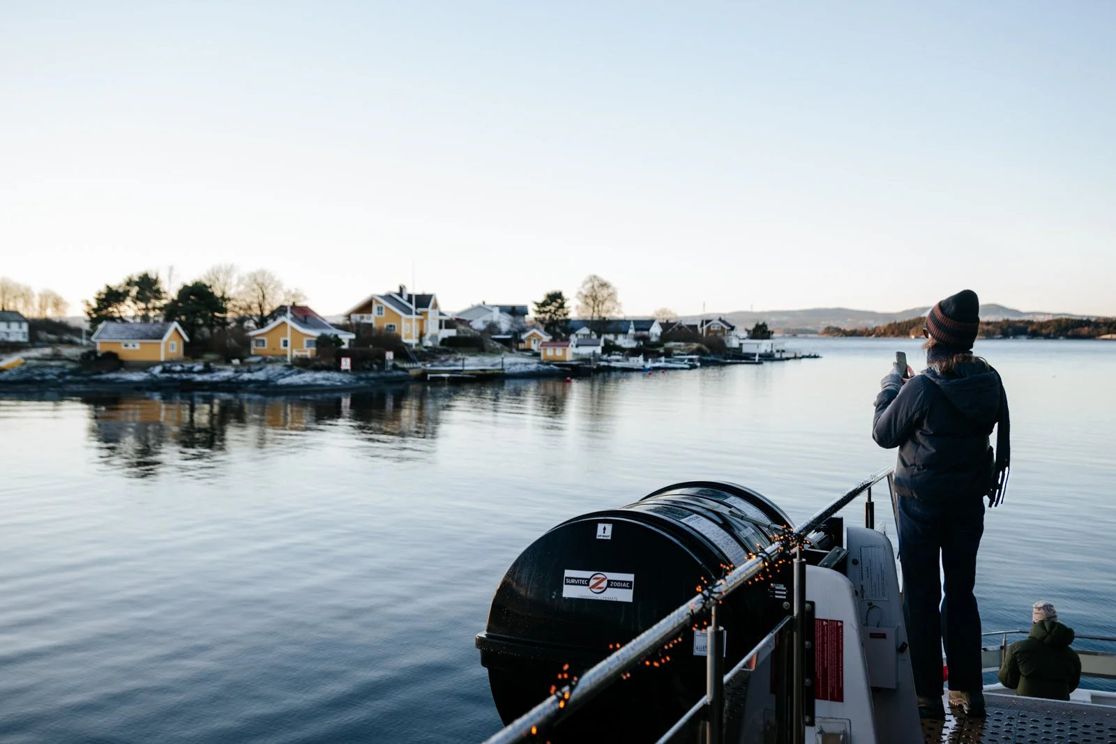 A person standing on a boat taking a photo of a coastal village with yellow houses along the water, with another person sitting on the boat, during a clear day.
