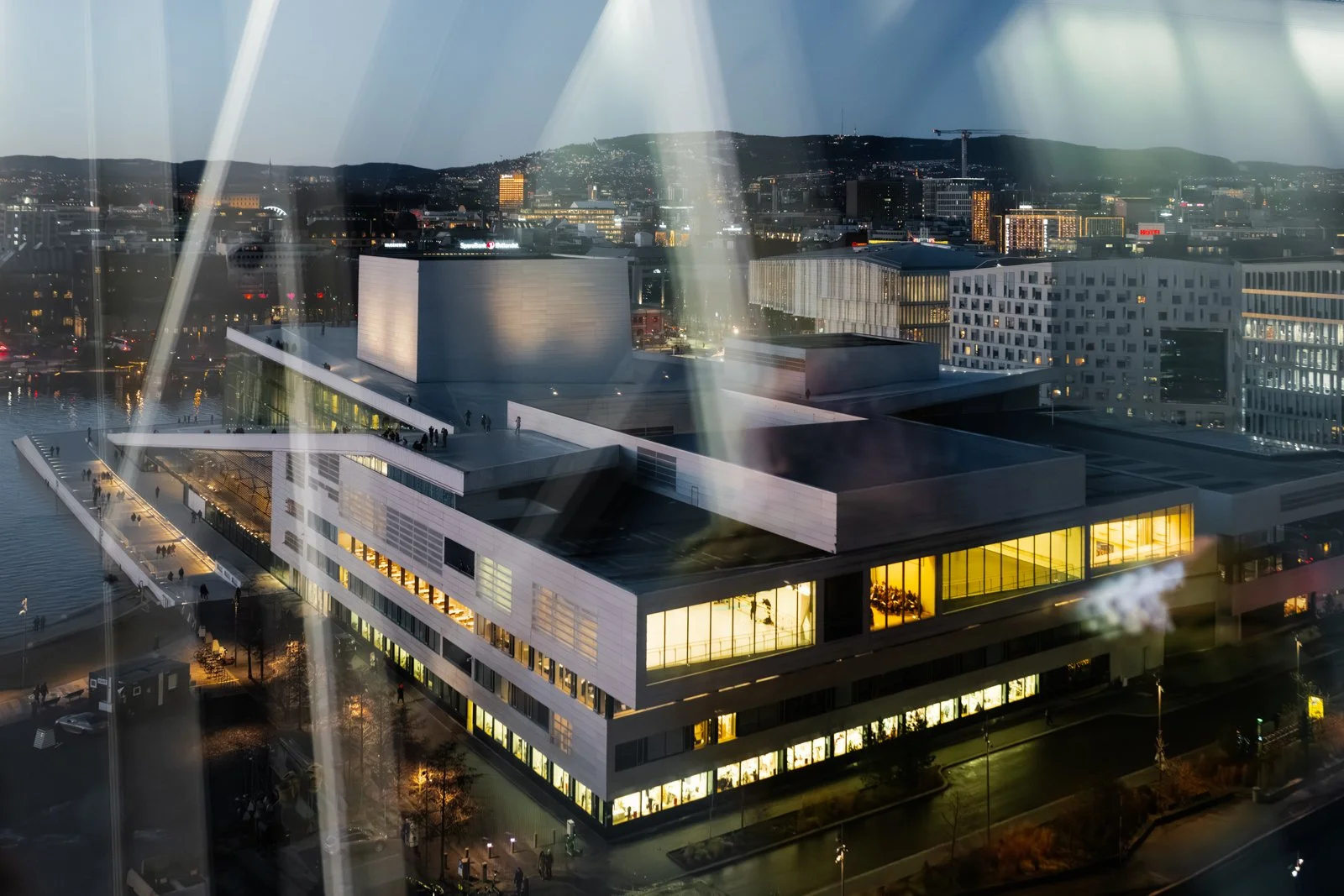 A cityscape at dusk with a modern multi-story building illuminated from within, seen through a glass window with reflections. The building is near a body of water, with other illuminated buildings and hills in the background.