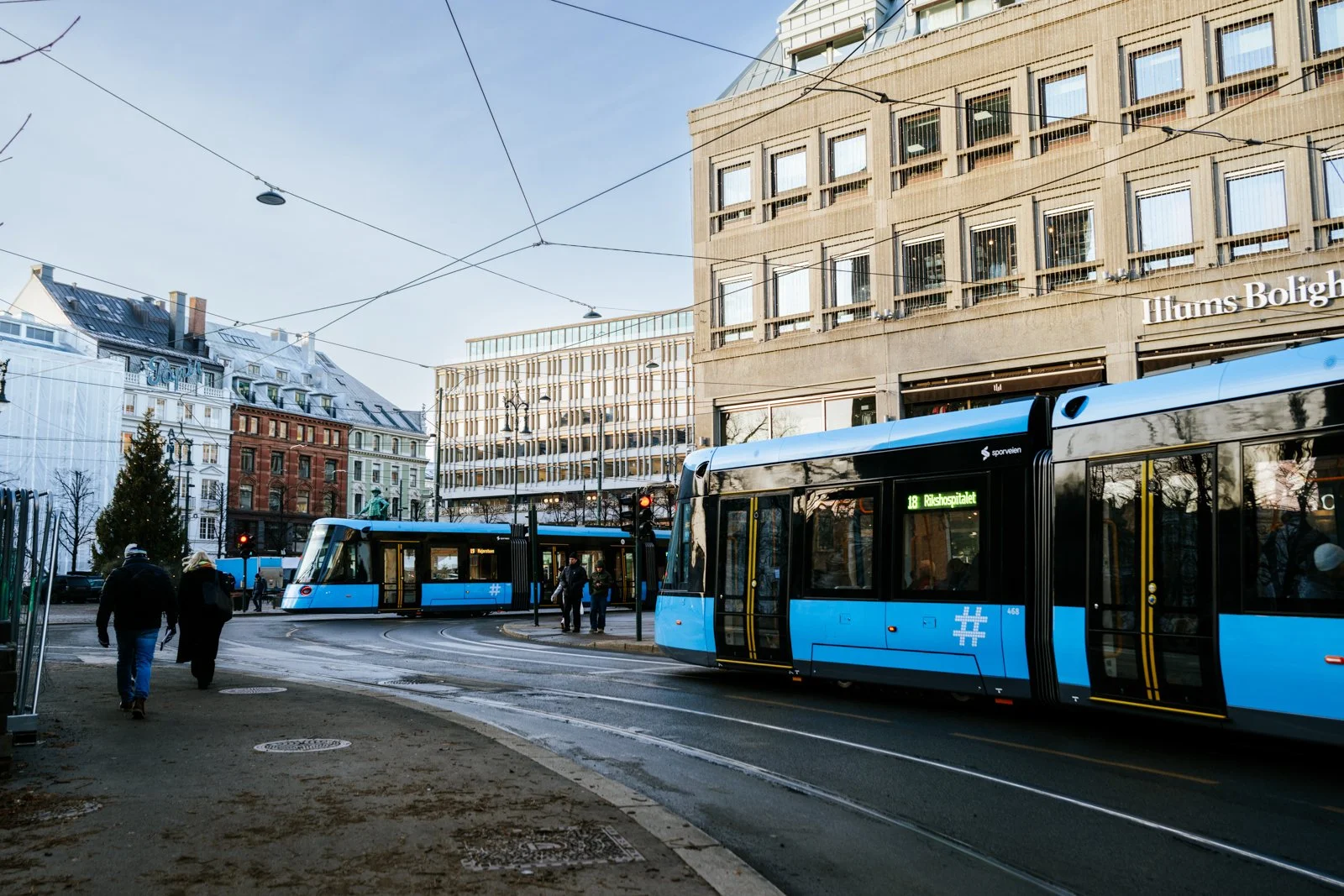 City street scene with two blue modern trams and pedestrians, buildings in the background, and clear weather.